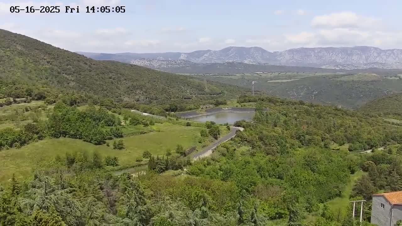 A partly cloudy day reveals a verdant landscape of rolling hills, lush green forests, a small reservoir, and a winding road, all set against a backdrop of distant mountains.