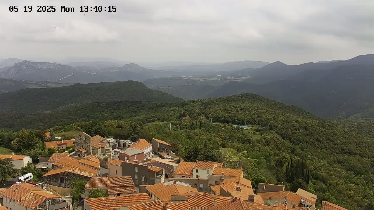 A village of orange-roofed buildings sits on a hillside overlooking a vast expanse of green mountains under a cloudy sky.