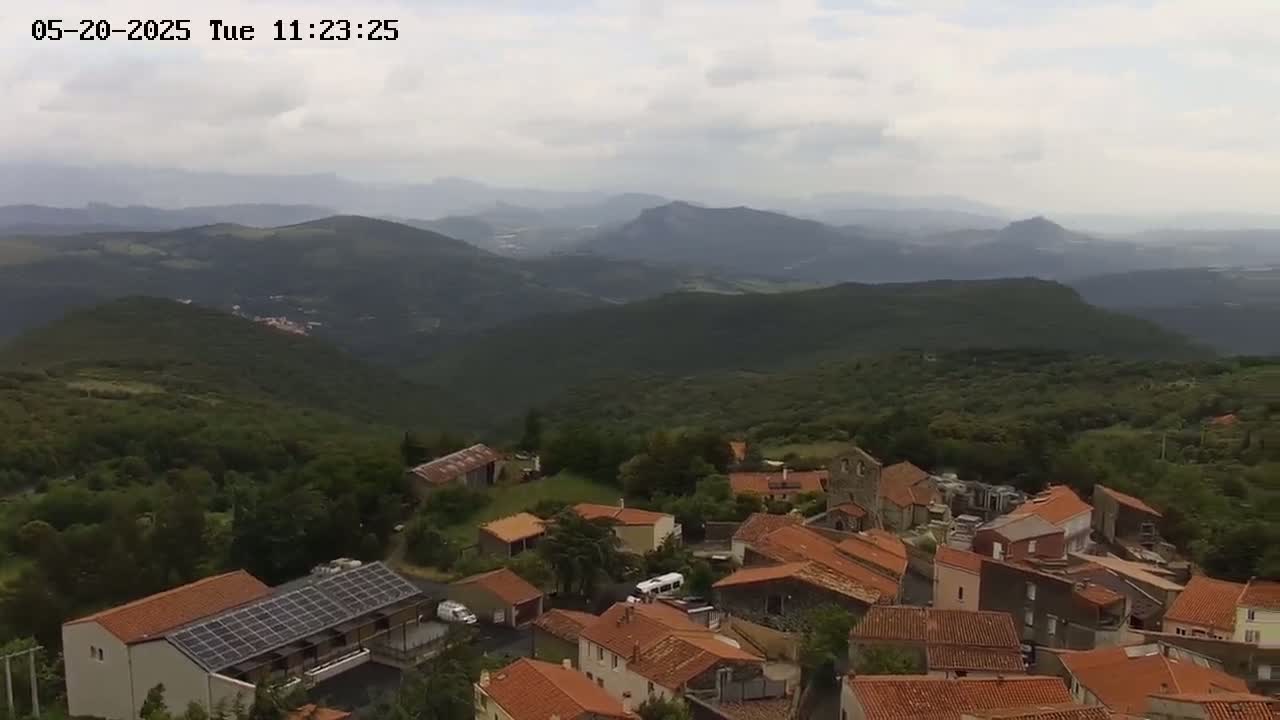 An aerial view shows a small village nestled in a mountainous region under a mostly cloudy sky.