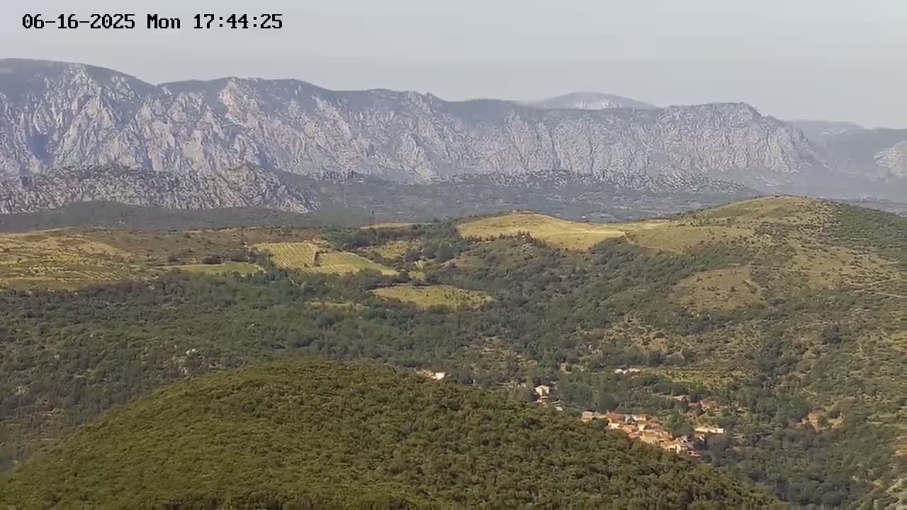 A hazy view shows a verdant valley dotted with small buildings and vineyards, nestled between rolling hills and a distant, rocky mountain range.