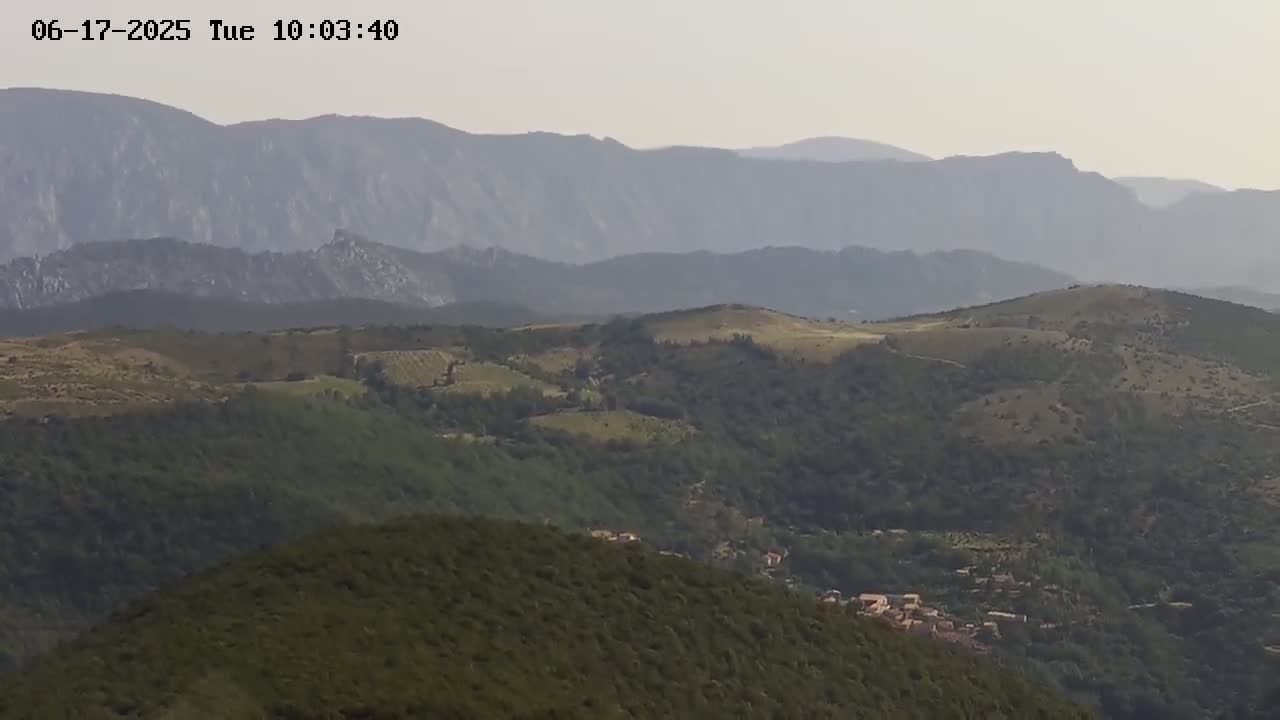 A hazy, distant mountain range overlooks rolling hills covered in green vegetation and a small village nestled in the valley below.