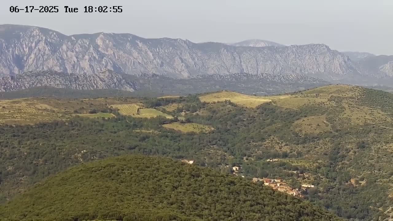 A hazy view shows rolling hills covered in green vegetation, with a small village nestled in the valley and a large mountain range in the background.