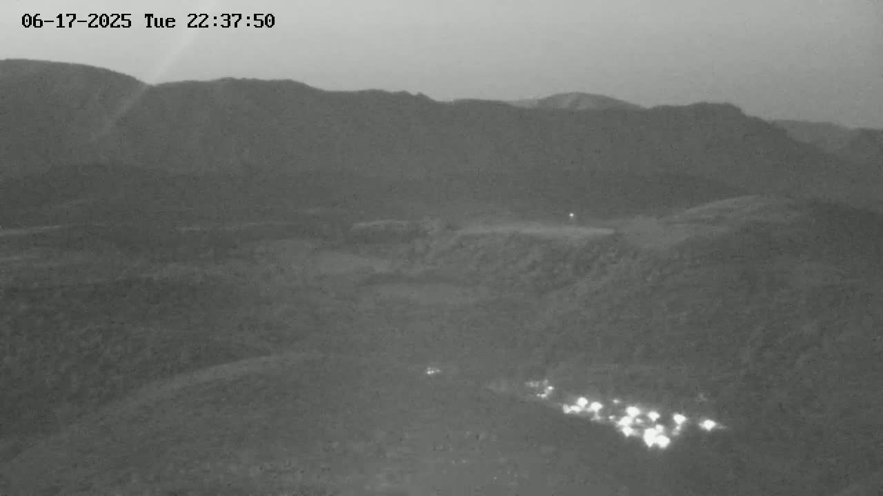A nighttime, low-light image shows a hilly, mountainous landscape with a line of headlights visible in the lower portion of the frame.