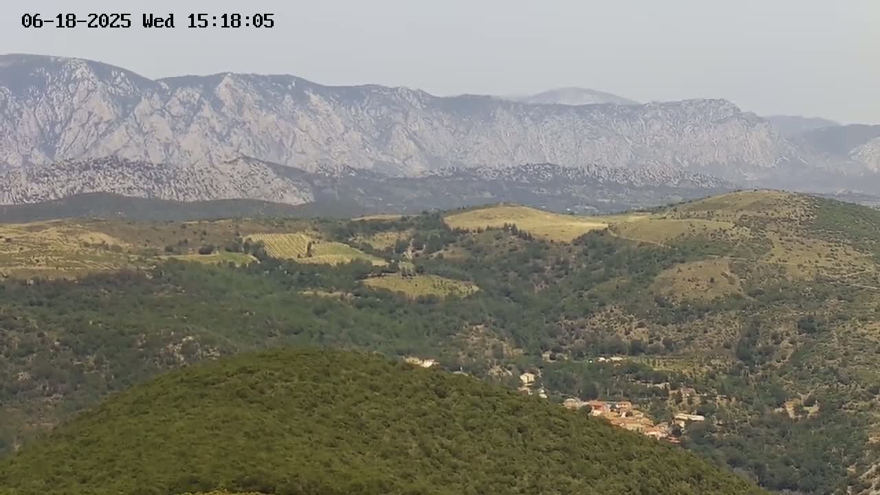 A hazy view of rolling hills and mountains covered in green vegetation, with some light-brown patches visible, under a mostly clear sky.