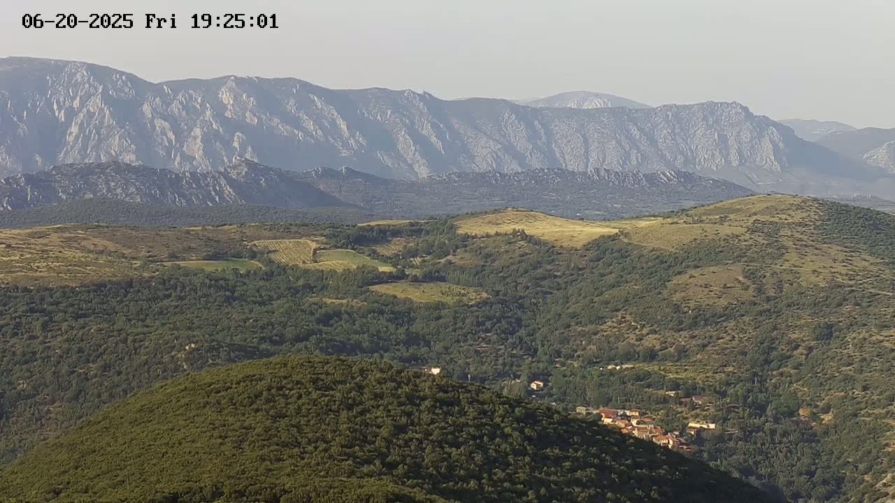 A vast hilly landscape, covered in green and brown vegetation, stretches towards a range of light-grey mountains under a hazy sky.