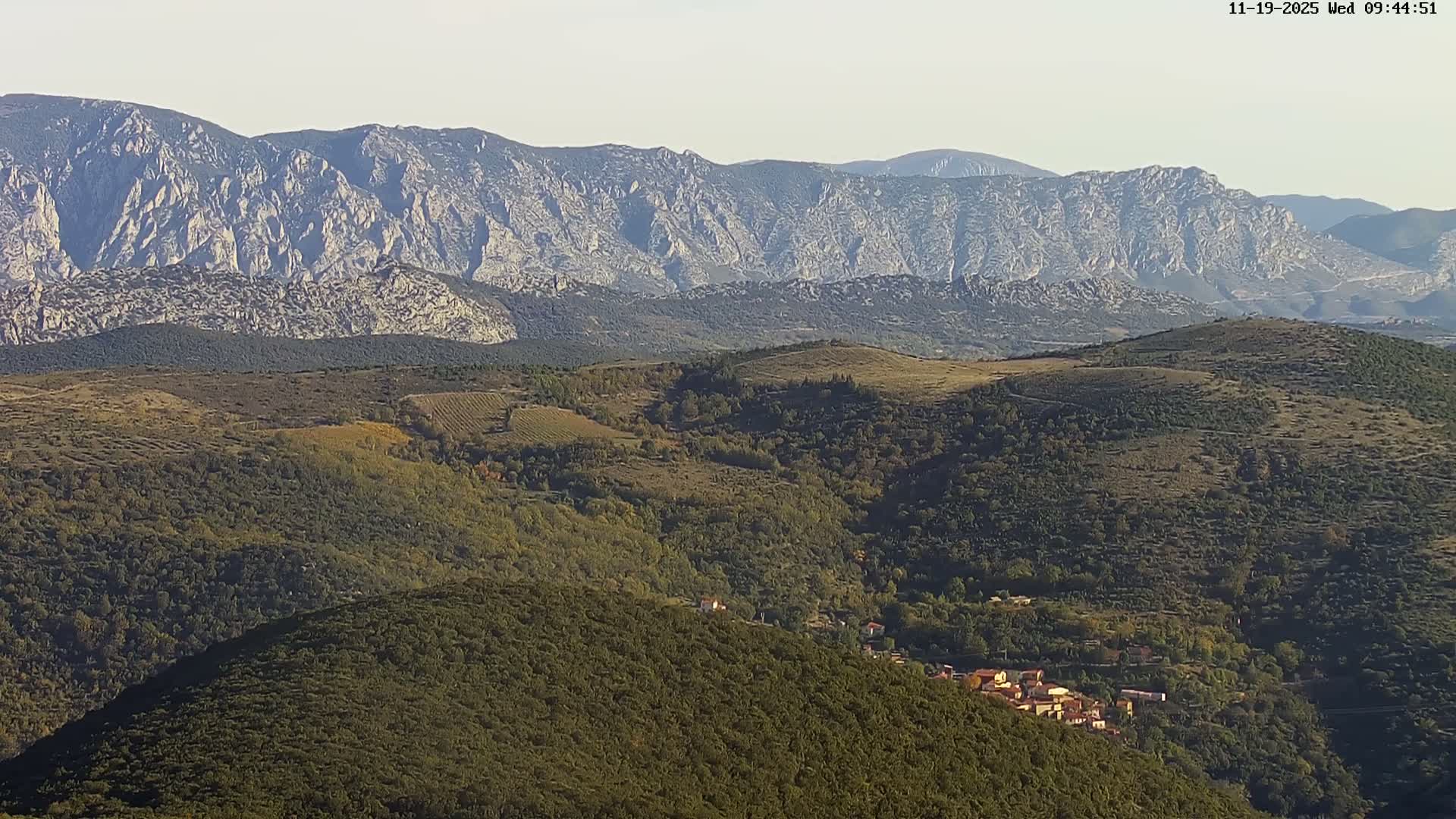 A clear, sunny day reveals a sprawling mountainous landscape with rugged peaks in the distance towering over densely forested hills and valleys, where a small, red-roofed village is nestled.