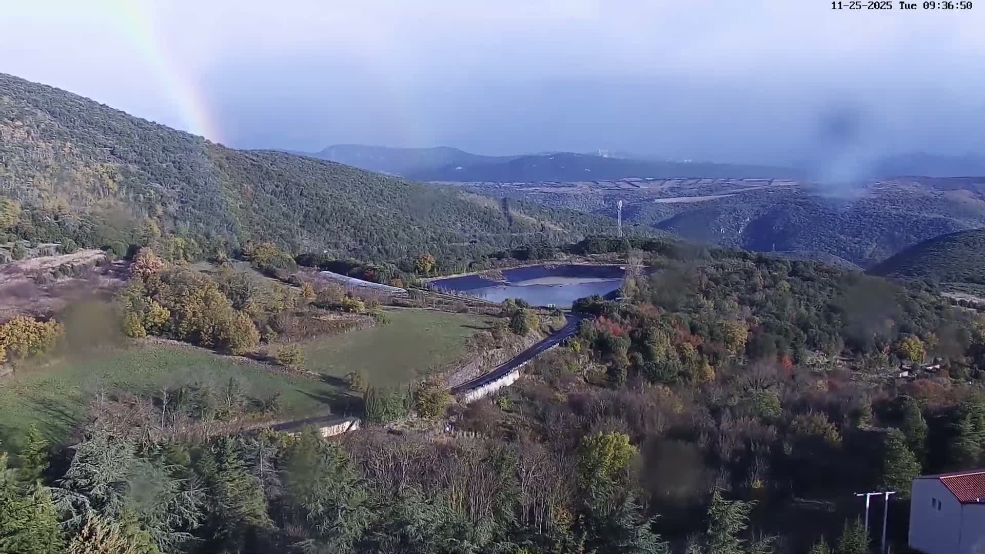 A panoramic view shows rolling hills covered in autumn foliage surrounding a reservoir and a road, with a vibrant rainbow arching across the sky on a bright, post-rain day.