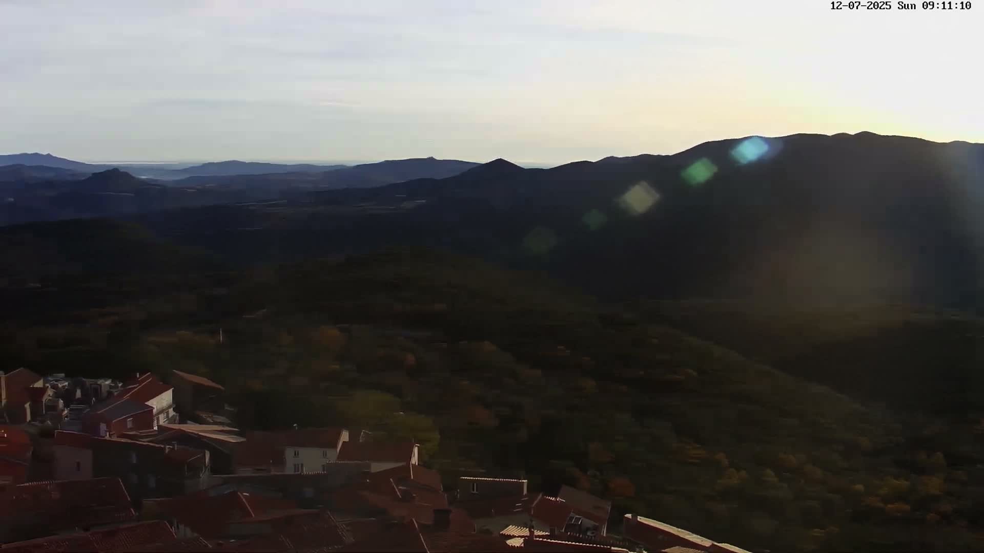 An expansive aerial view reveals a village with red-tiled roofs nestled below dark, rolling mountains and a distant sea, all illuminated by clear morning sunlight with visible lens flares.