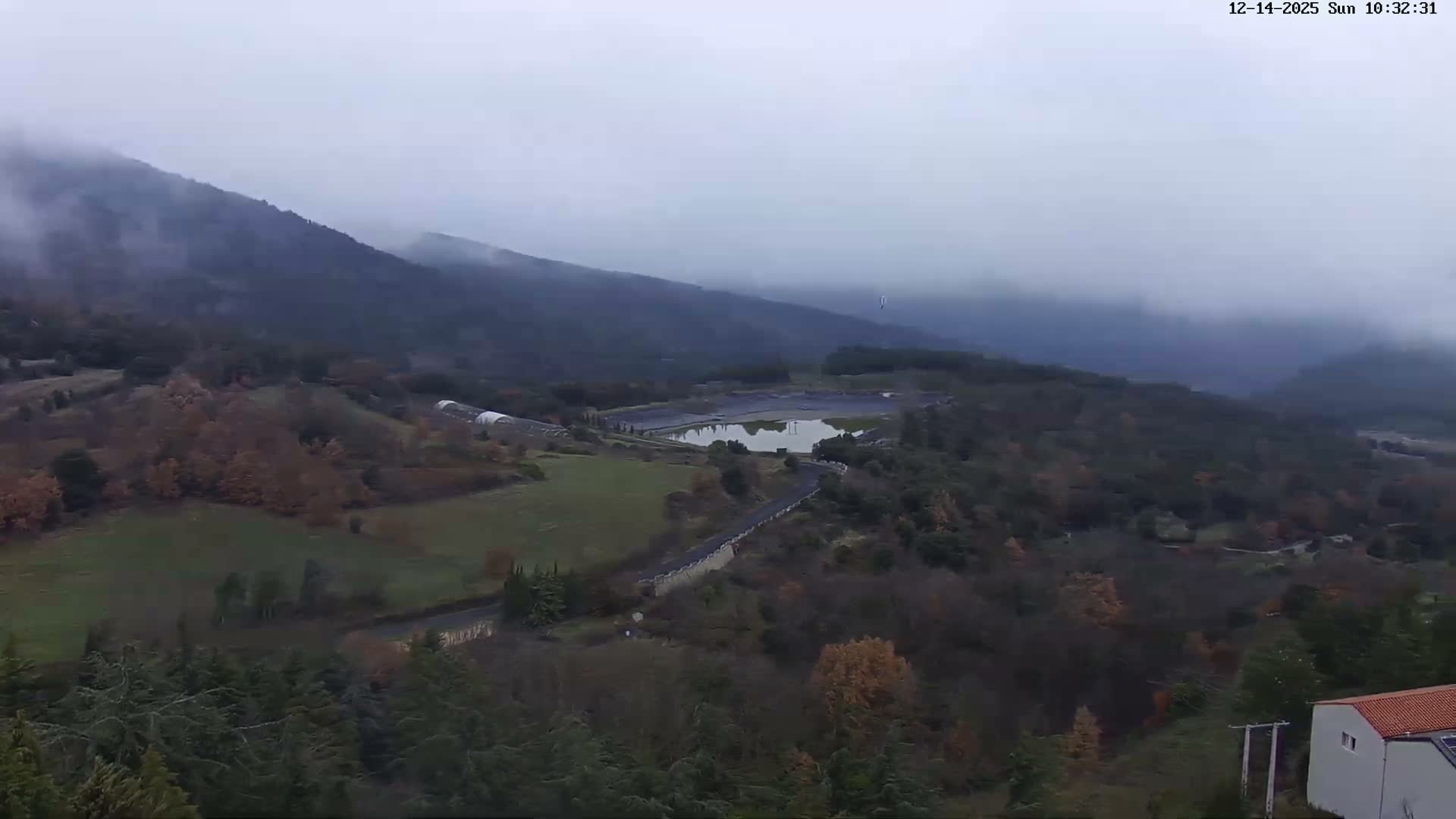An expansive aerial view reveals a village with red-tiled roofs nestled below dark, rolling mountains and a distant sea, all illuminated by clear morning sunlight with visible lens flares.