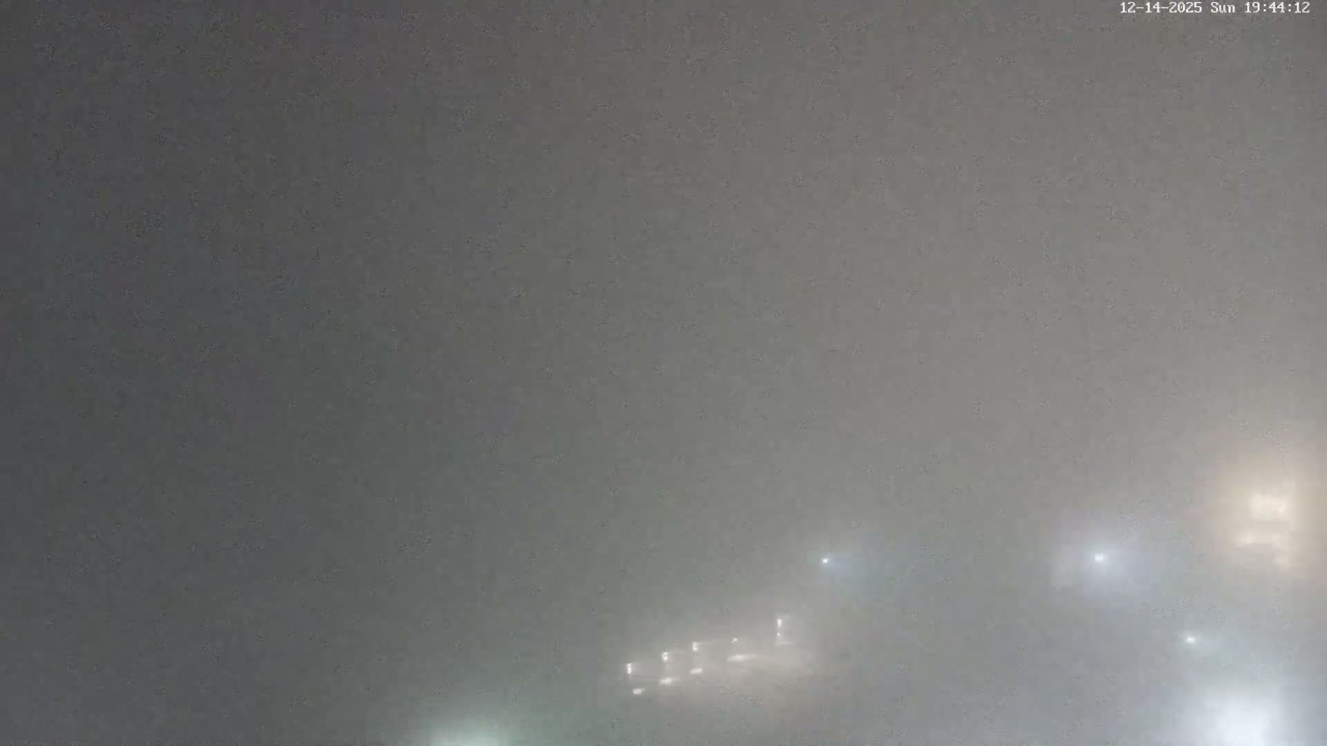 An expansive aerial view reveals a village with red-tiled roofs nestled below dark, rolling mountains and a distant sea, all illuminated by clear morning sunlight with visible lens flares.