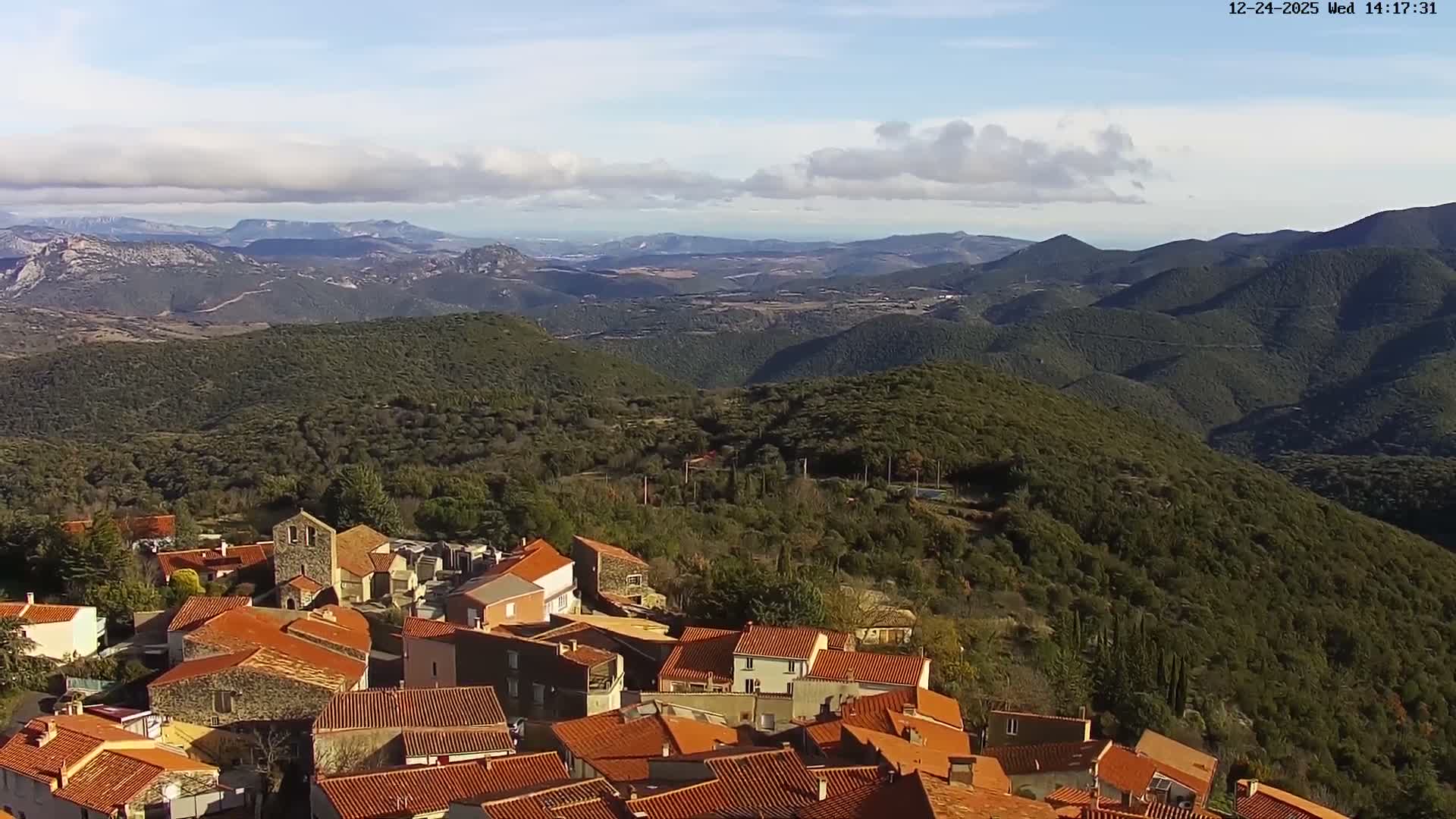 An expansive aerial view reveals a village with red-tiled roofs nestled below dark, rolling mountains and a distant sea, all illuminated by clear morning sunlight with visible lens flares.