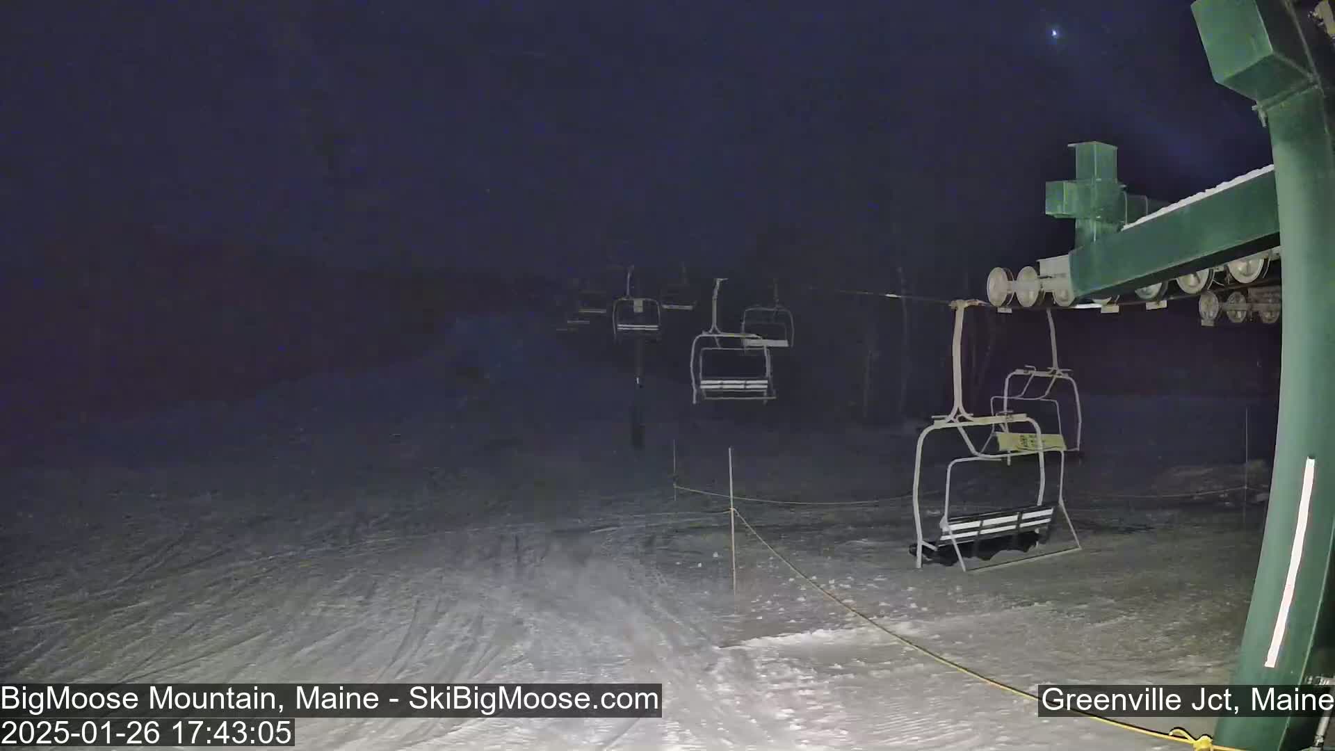 Under a dark, nighttime sky, empty ski lift chairs hang from cables above a snow-covered slope.