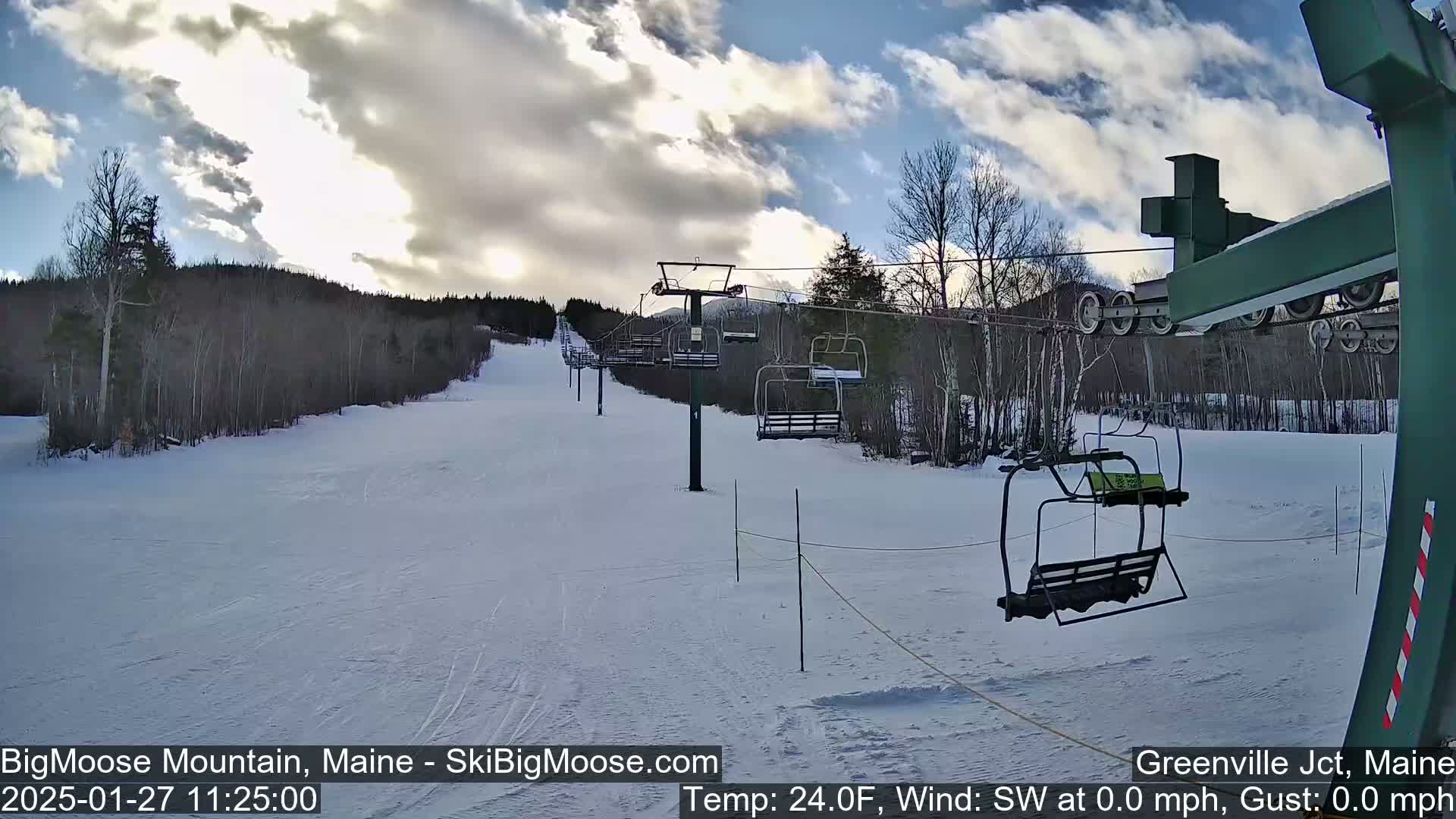 A mostly sunny, snow-covered ski slope with a chairlift and sparsely wooded hills in the background.