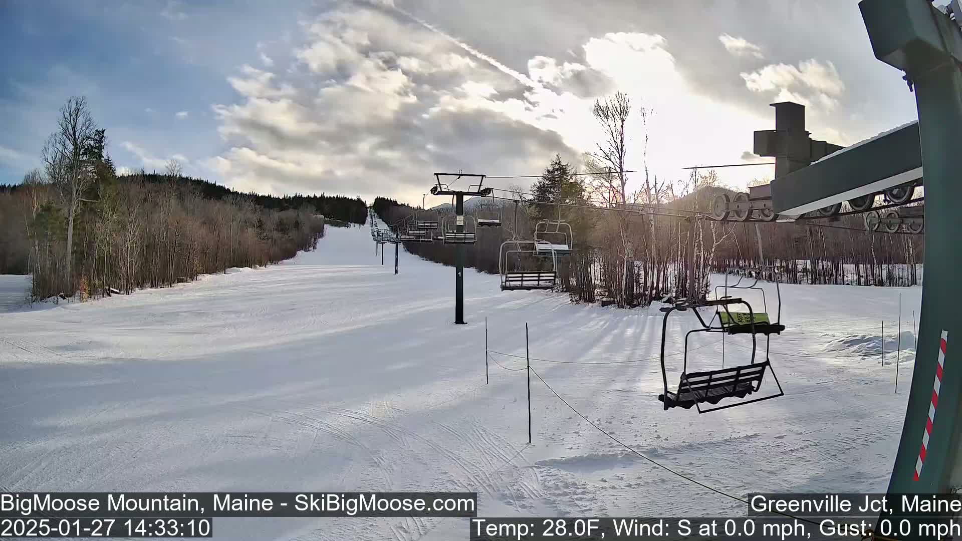 A snow-covered ski slope with a chairlift and sparsely wooded areas under a partly cloudy sky.