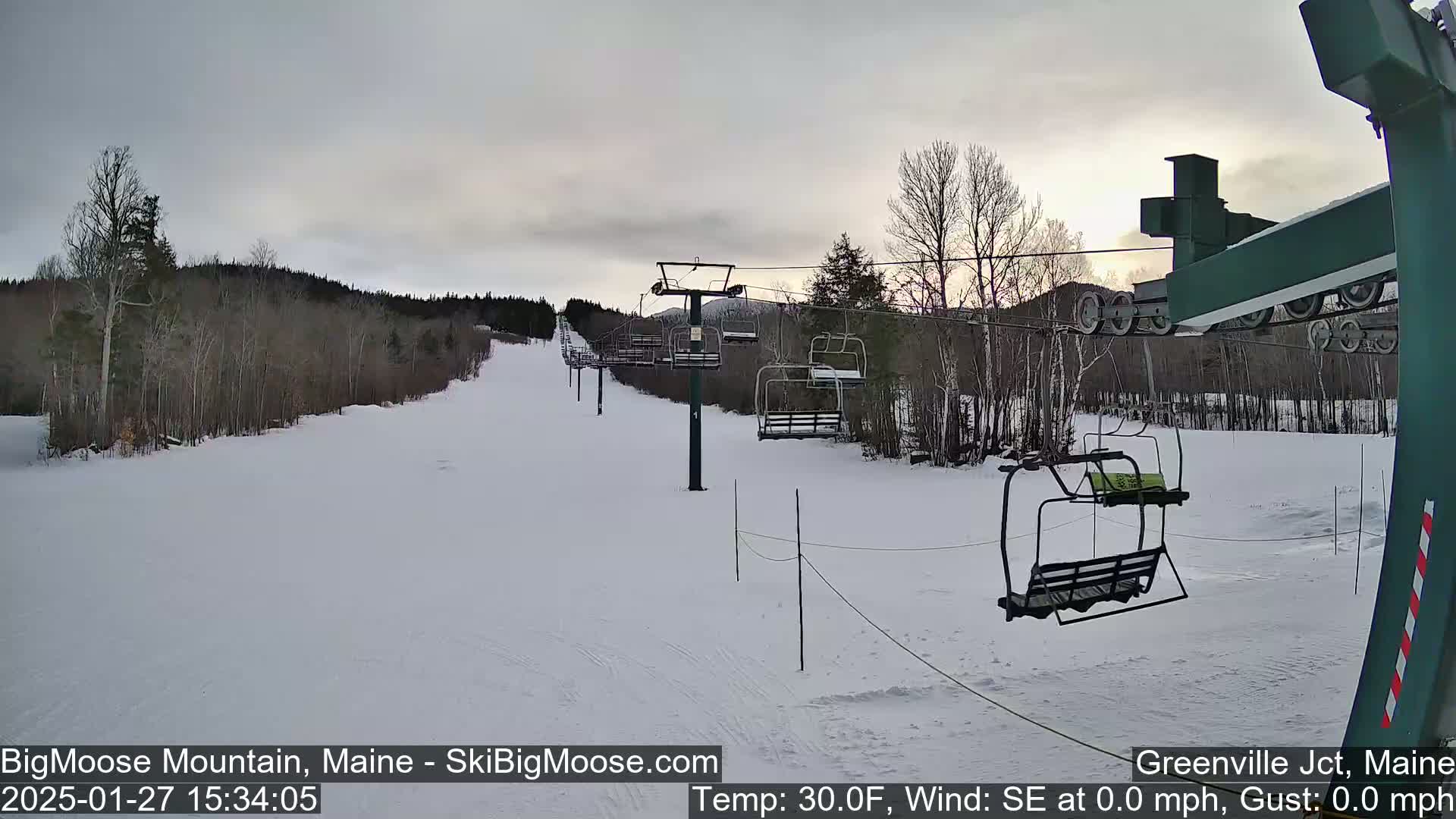 A snow-covered ski slope with a chairlift and sparsely wooded hills under an overcast sky.