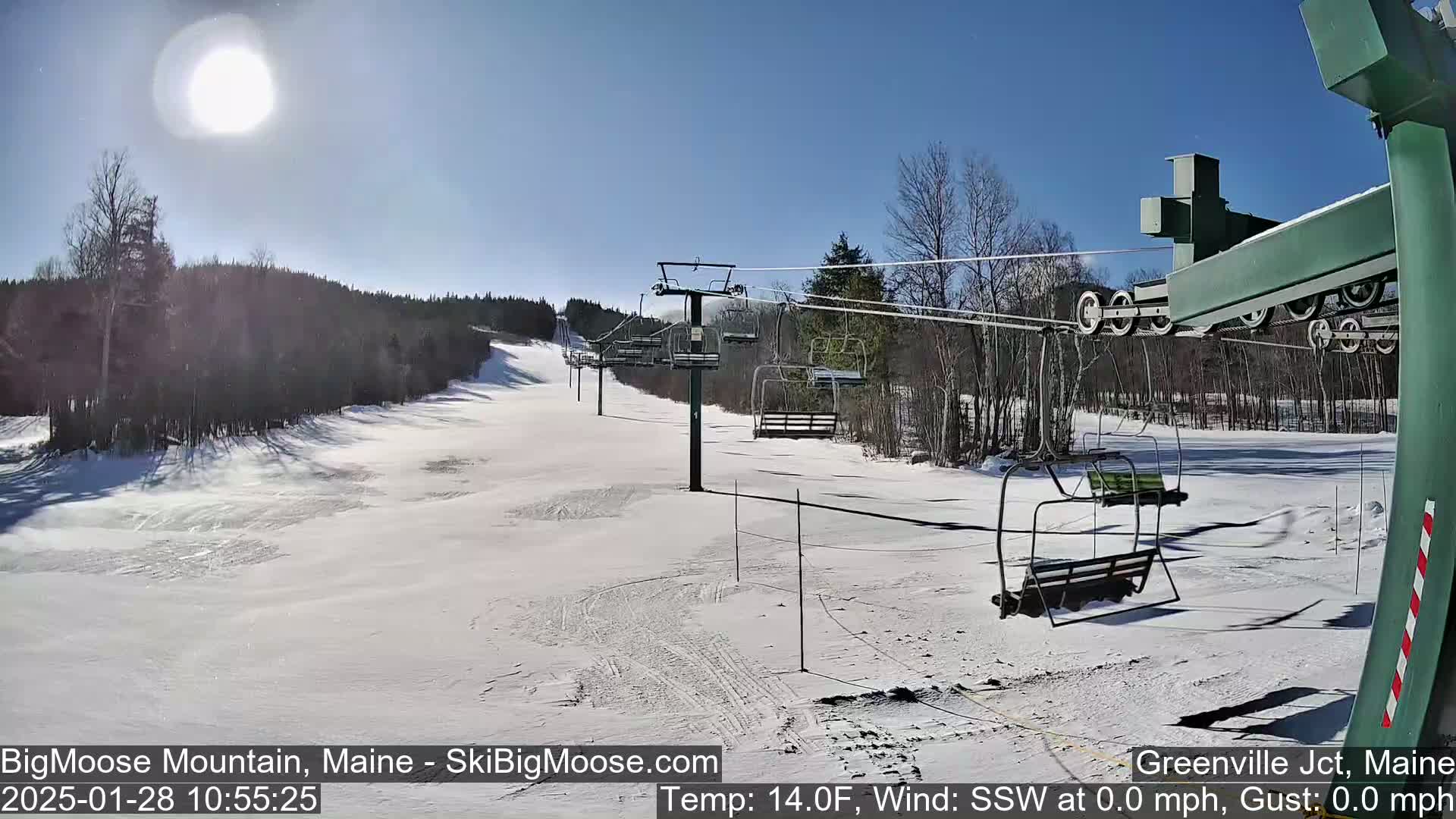 A sunny day at a ski resort shows a snow-covered slope with a chairlift and surrounding trees.