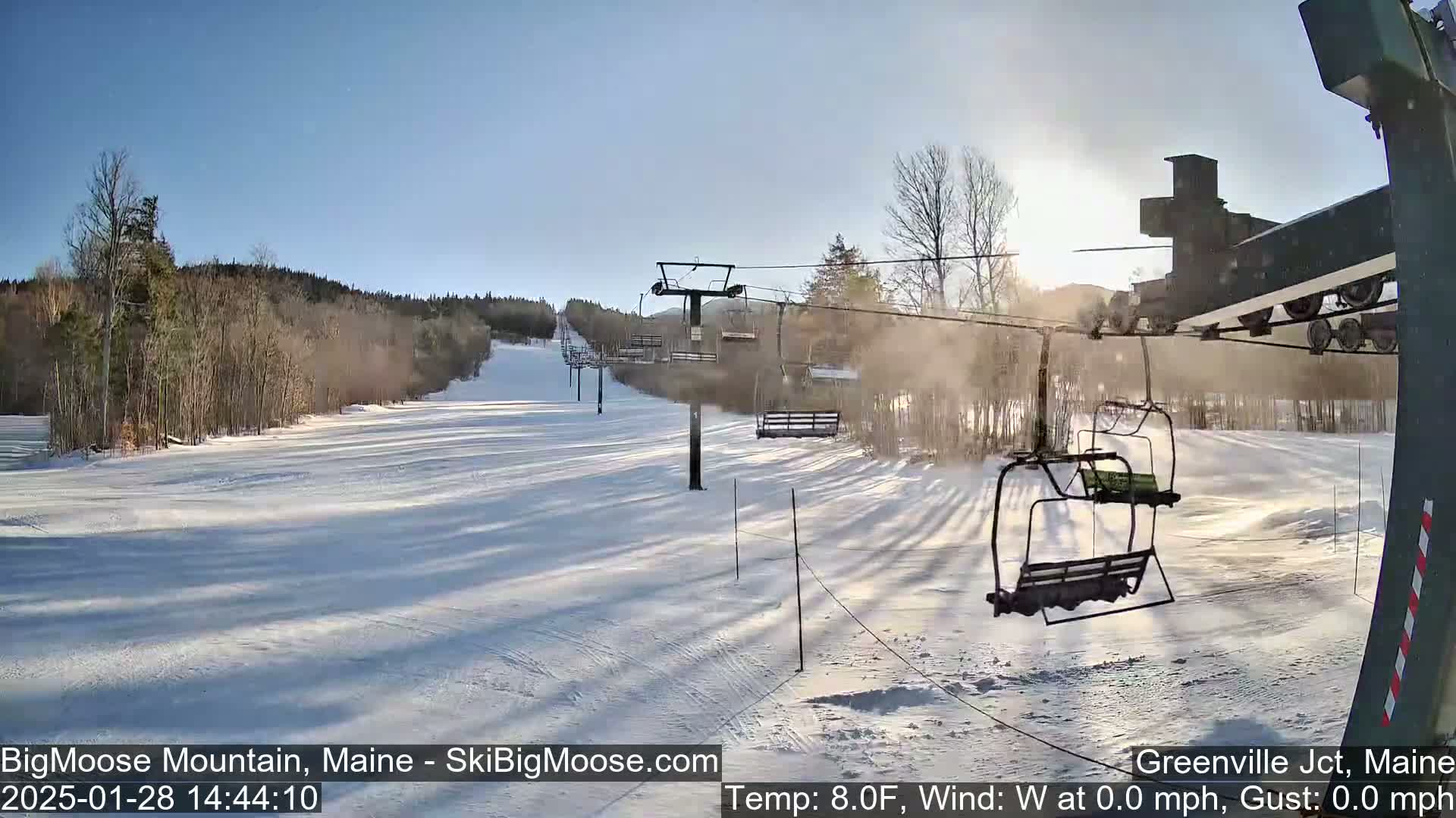 A sunny, snowy ski slope with a chairlift and empty chairs visible, under a clear blue sky.