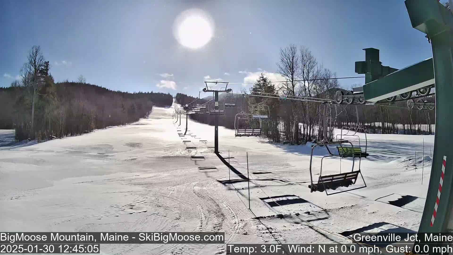 A sunny, snow-covered ski slope with a chairlift extends into a wooded area.