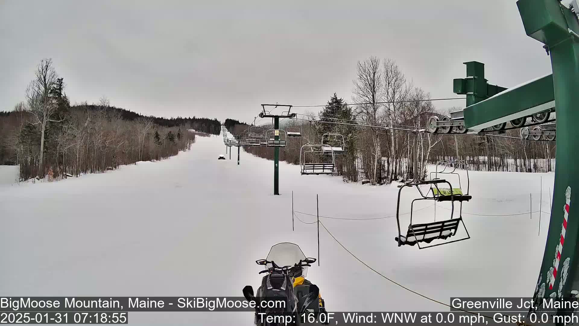 A snowmobile is positioned on a snowy ski slope beneath a chairlift, with bare trees lining the hills under an overcast sky.