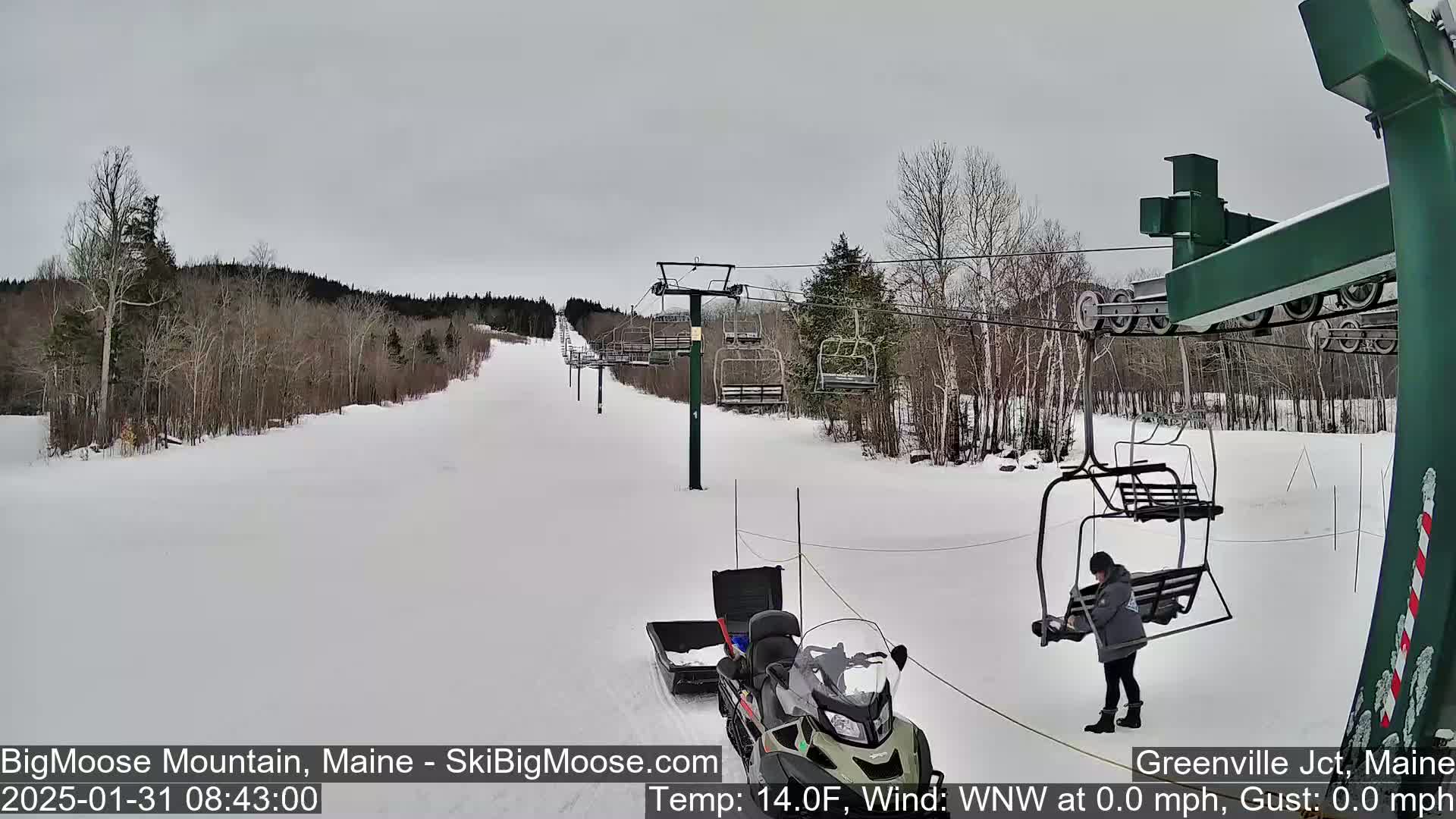 A snow-covered ski slope with a chairlift, a snowmobile, and a person standing near a chair.