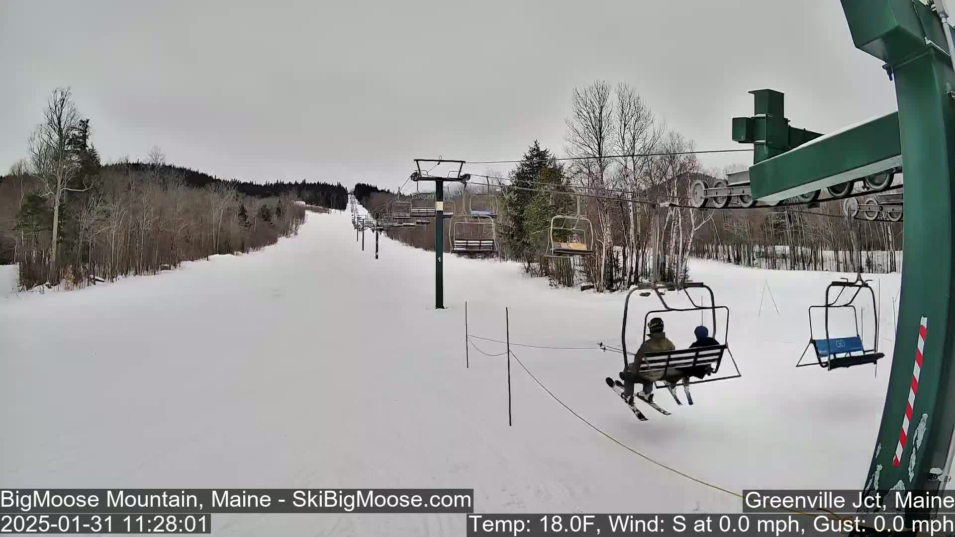 A chairlift ascends a snow-covered ski slope on an overcast day, carrying two skiers.