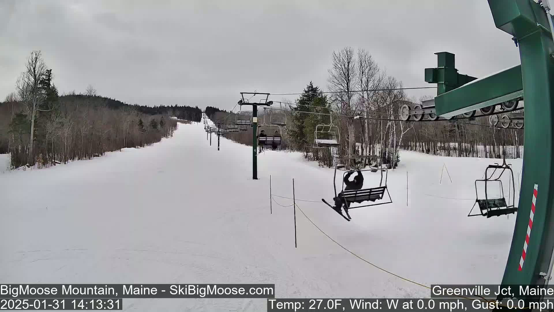 A ski lift ascends a snow-covered slope, with several skiers visible on the chairs against an overcast sky.