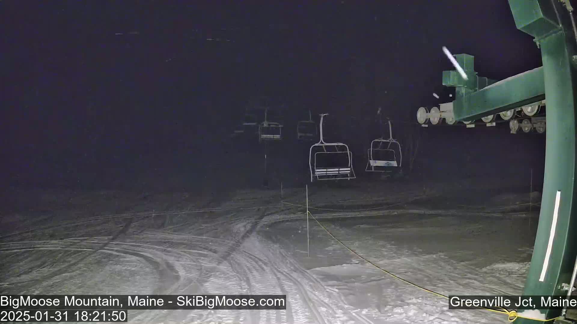 A nighttime, snowy scene shows several empty chairlifts at a ski resort under light snowfall.