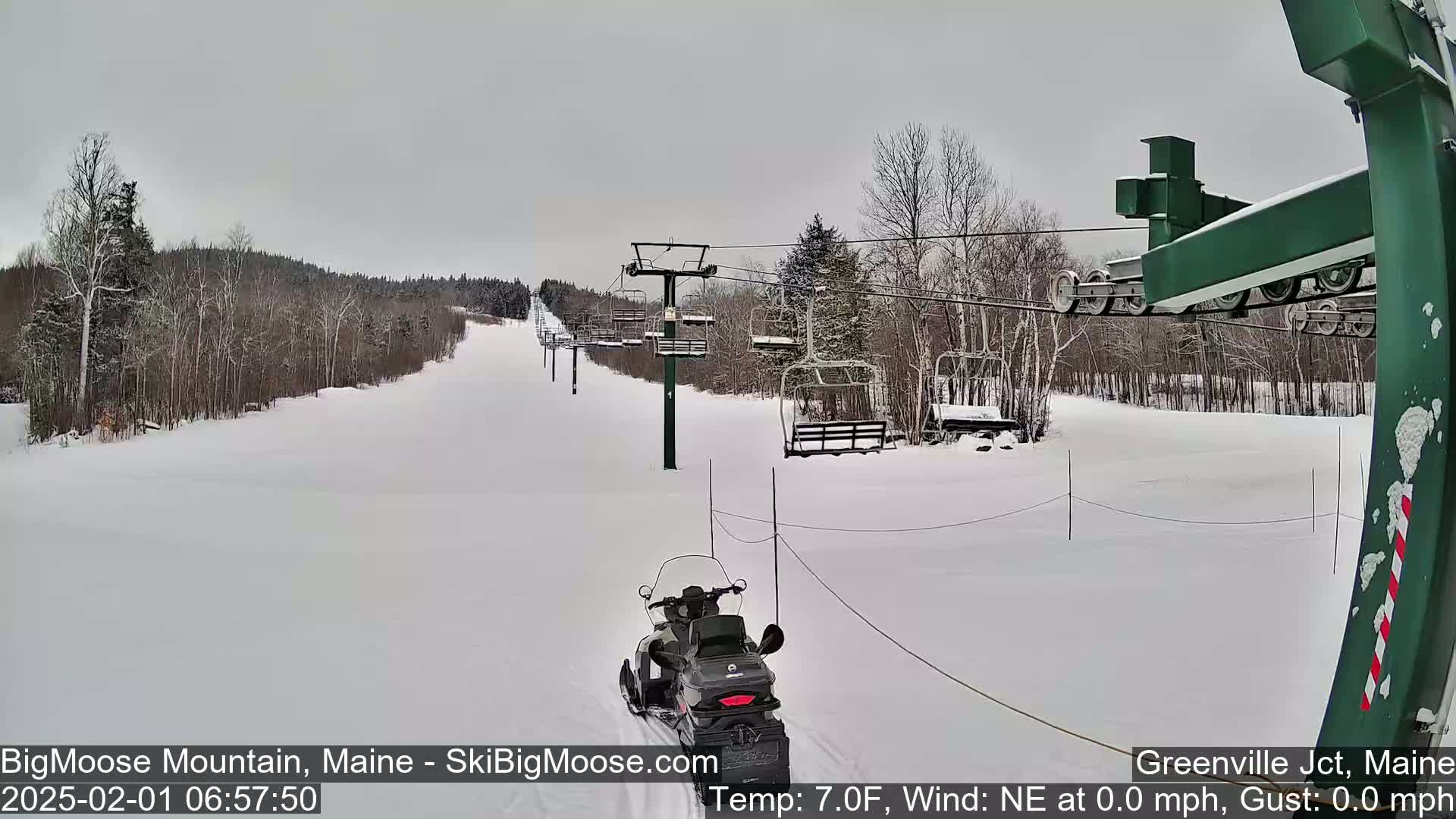A snowmobile travels up a snow-covered ski slope toward a chairlift under an overcast sky.