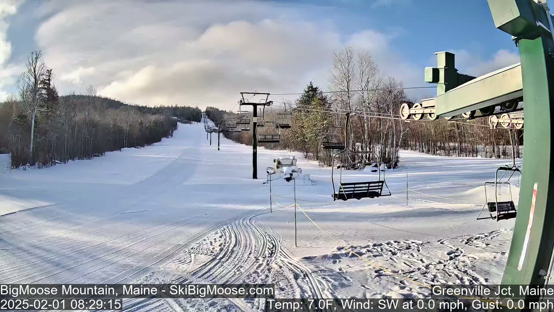 A snow-covered ski slope with several chairlifts and a groomed trail under a partly cloudy sky.