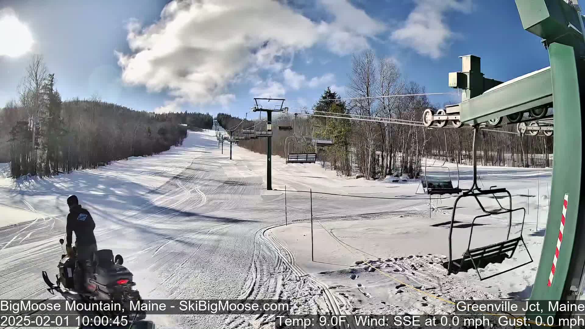 A snowmobile travels on a snow-covered ski slope under a partly cloudy sky, with a chairlift visible in the background.