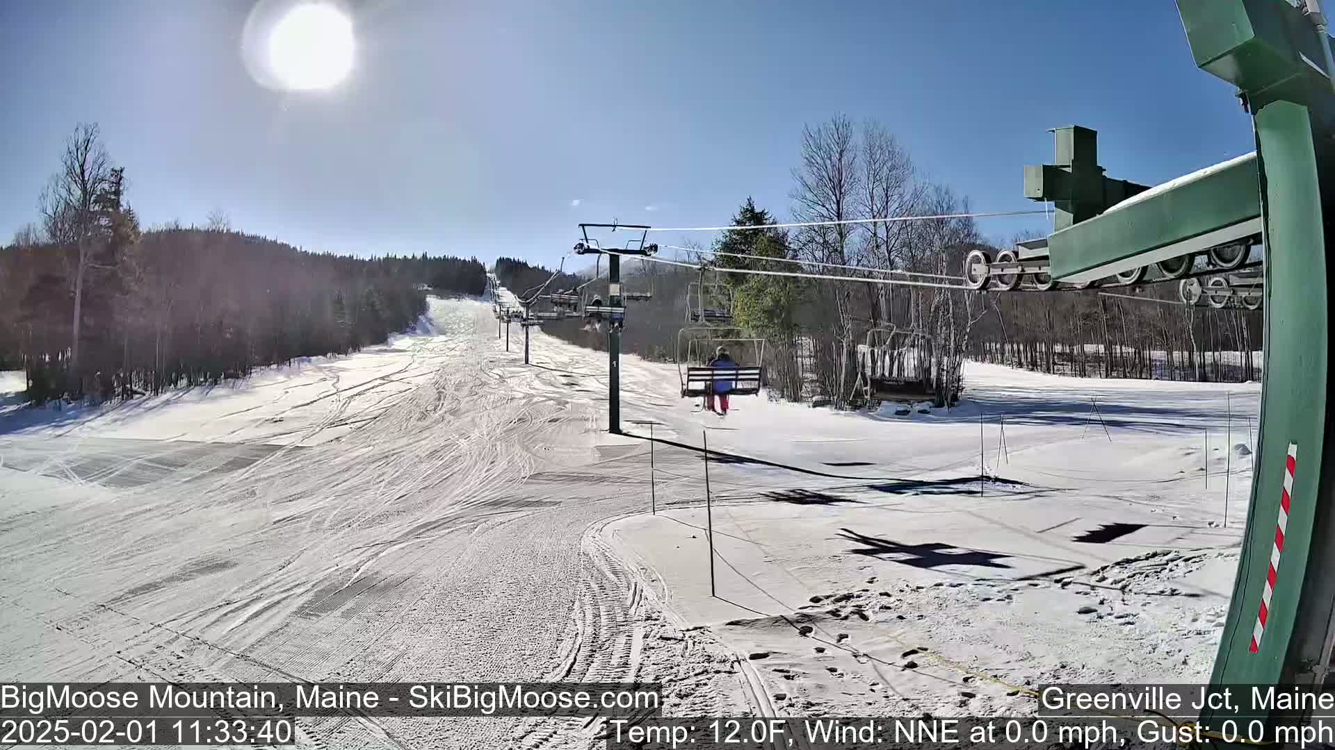 A sunny, snow-covered ski slope with a chairlift and a skier visible on the lift.