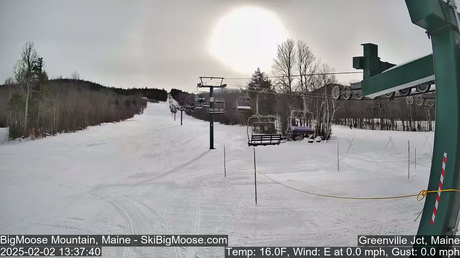 A mostly sunny, cold day on a snow-covered ski slope with a chairlift and sparsely wooded hills in the background.