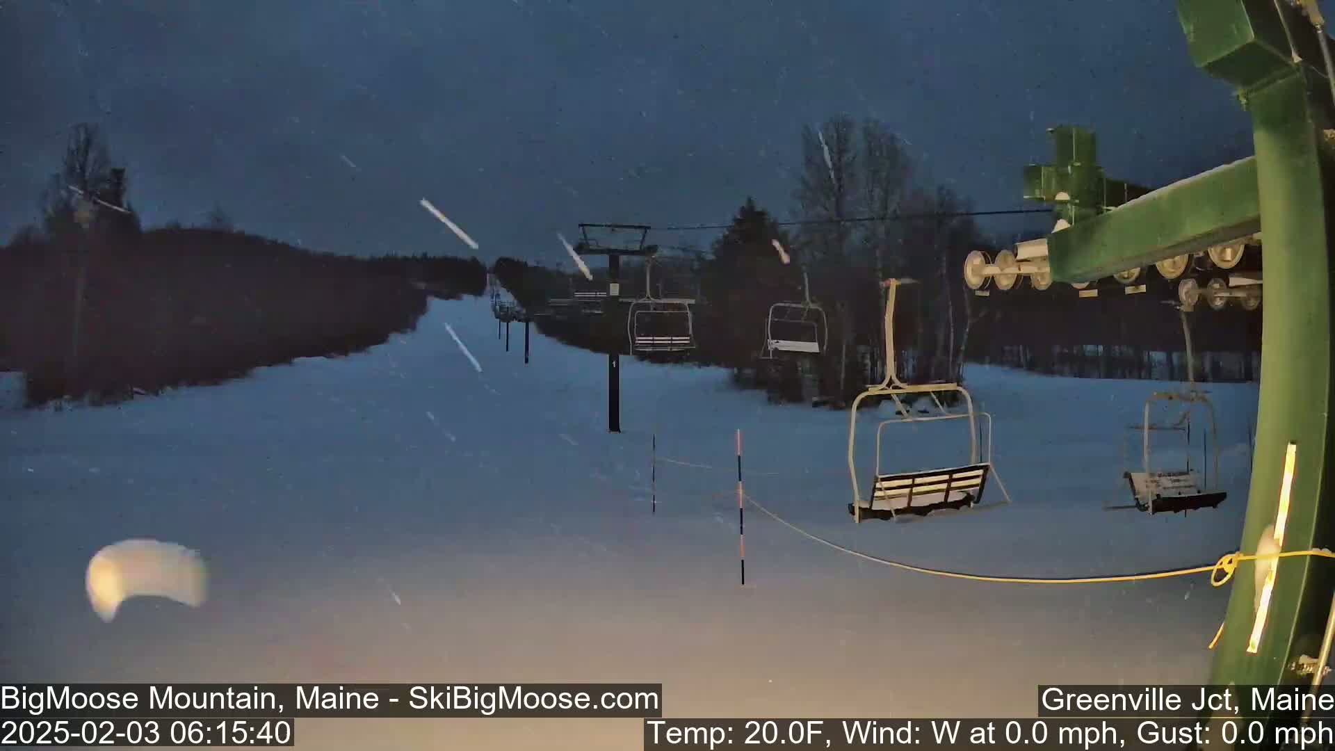 A snow-covered ski lift with empty chairs is visible under a dark, nighttime sky with light snowfall.