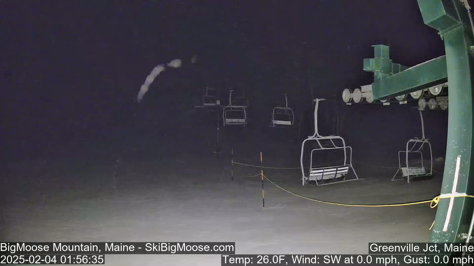 A nighttime snow scene shows empty ski lift chairs on a snow-covered slope, with light snowfall.