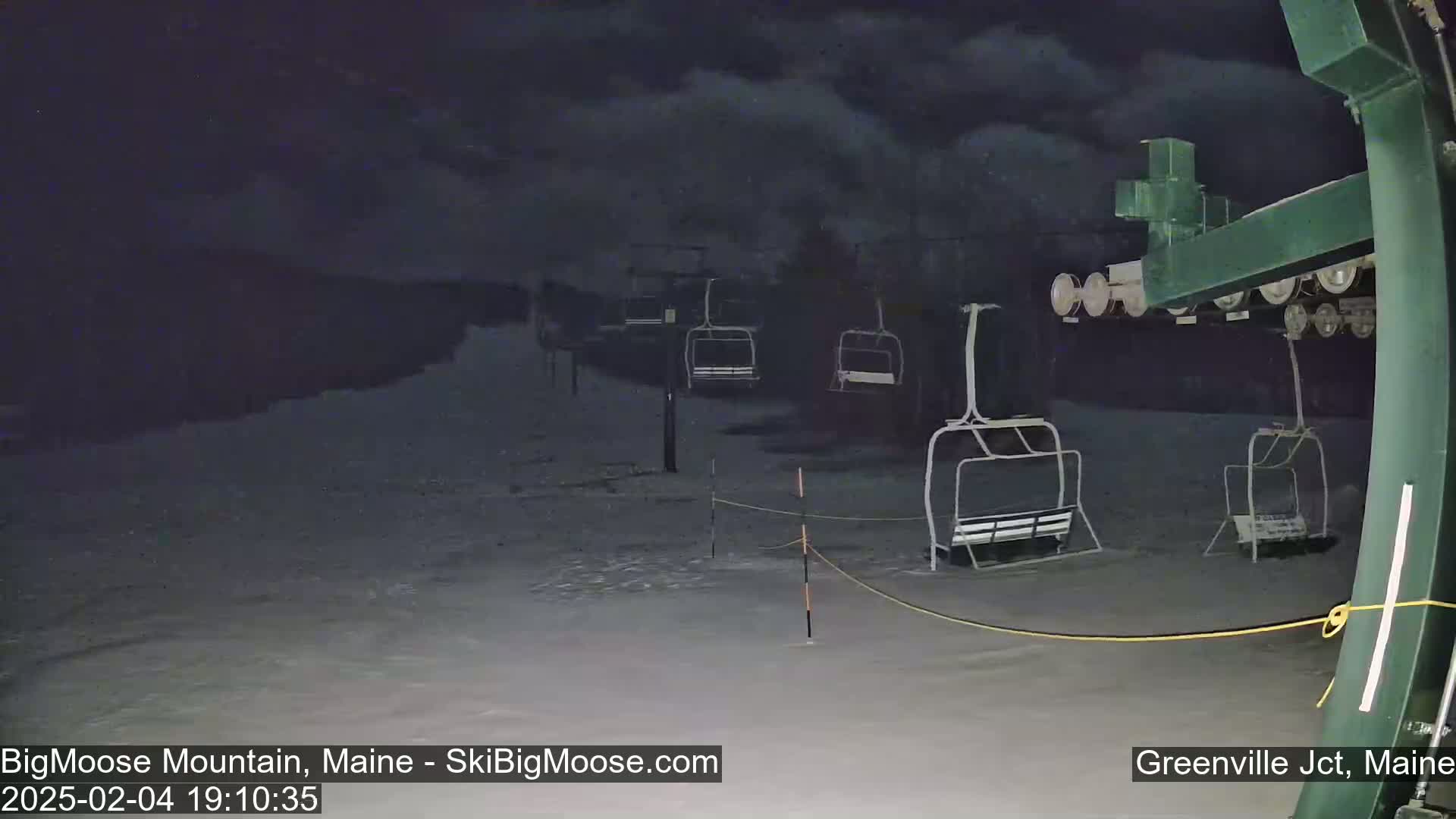 A nighttime view of a snow-covered ski lift with several empty chairlifts under a dark, cloudy sky.