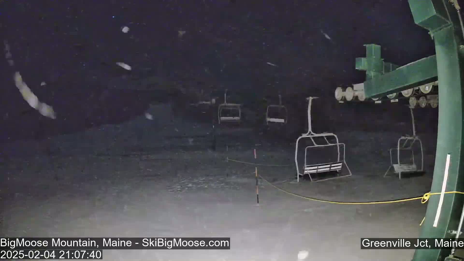 Several empty chairlifts sit on a snowy, dark ski slope at night under falling snow.
