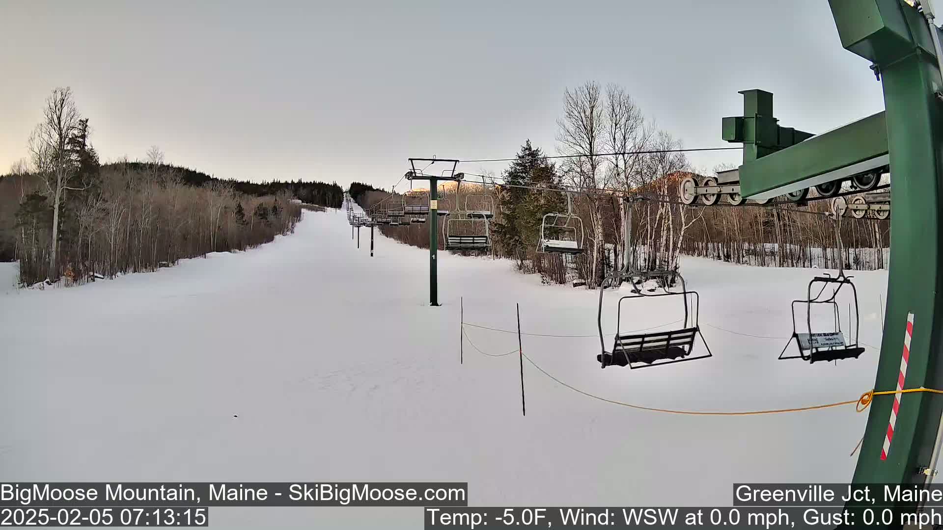 A snow-covered ski slope with an empty chairlift extends toward a line of trees under a pale gray sky.