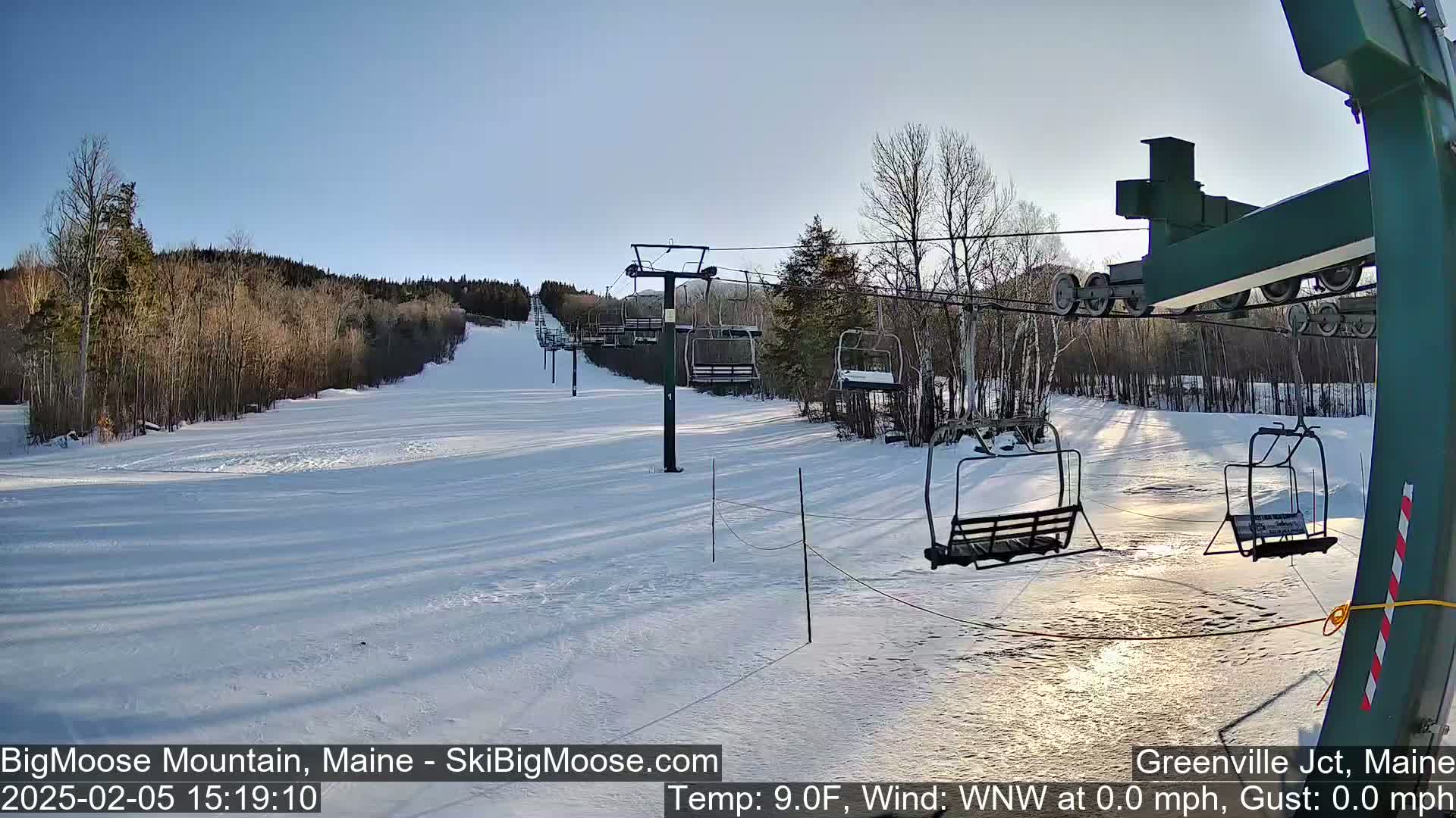 A snow-covered ski slope with a chairlift and surrounding trees under a clear, sunny sky.