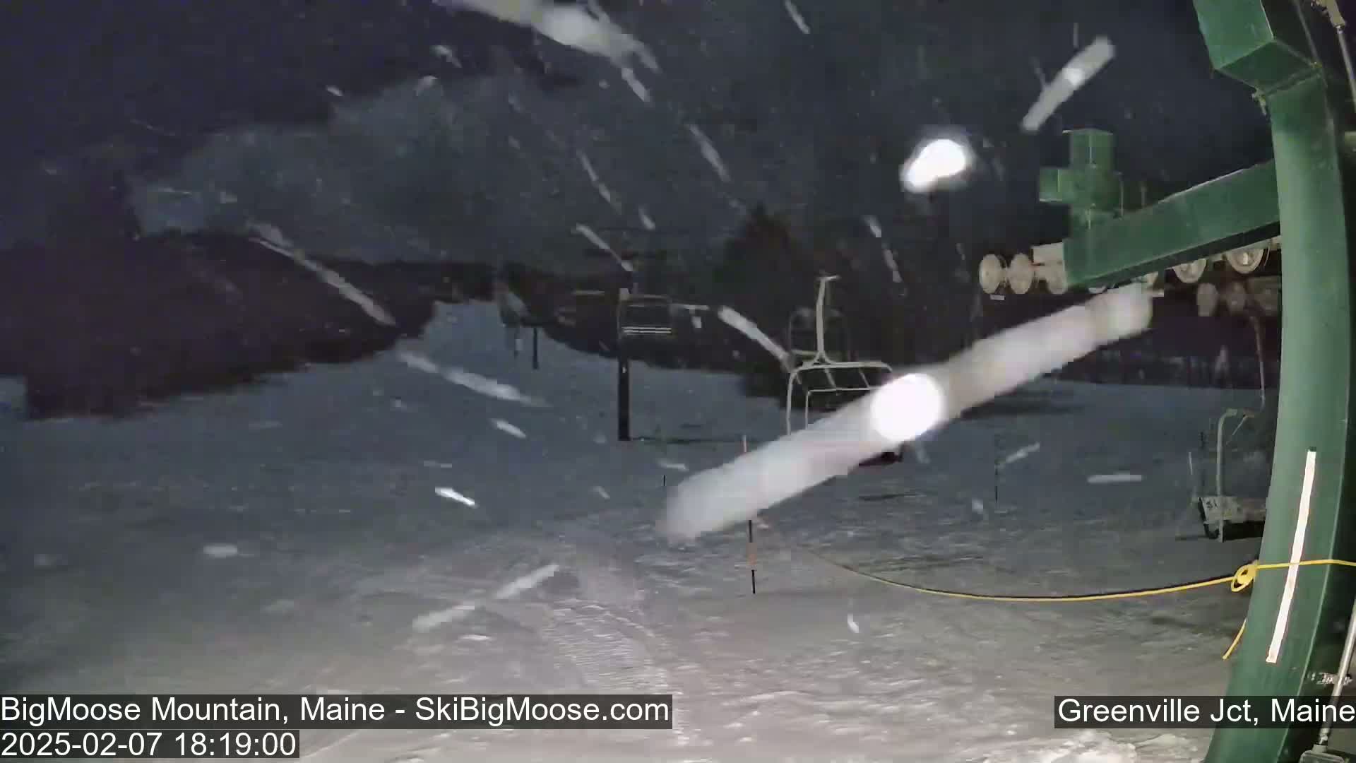A snow-covered ski slope at night is seen through a ski lift, with heavy snowfall actively occurring.