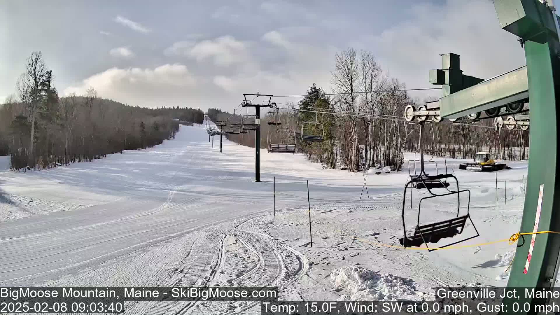 A snow-covered ski slope with a chairlift and sparsely wooded area under a partly cloudy sky.