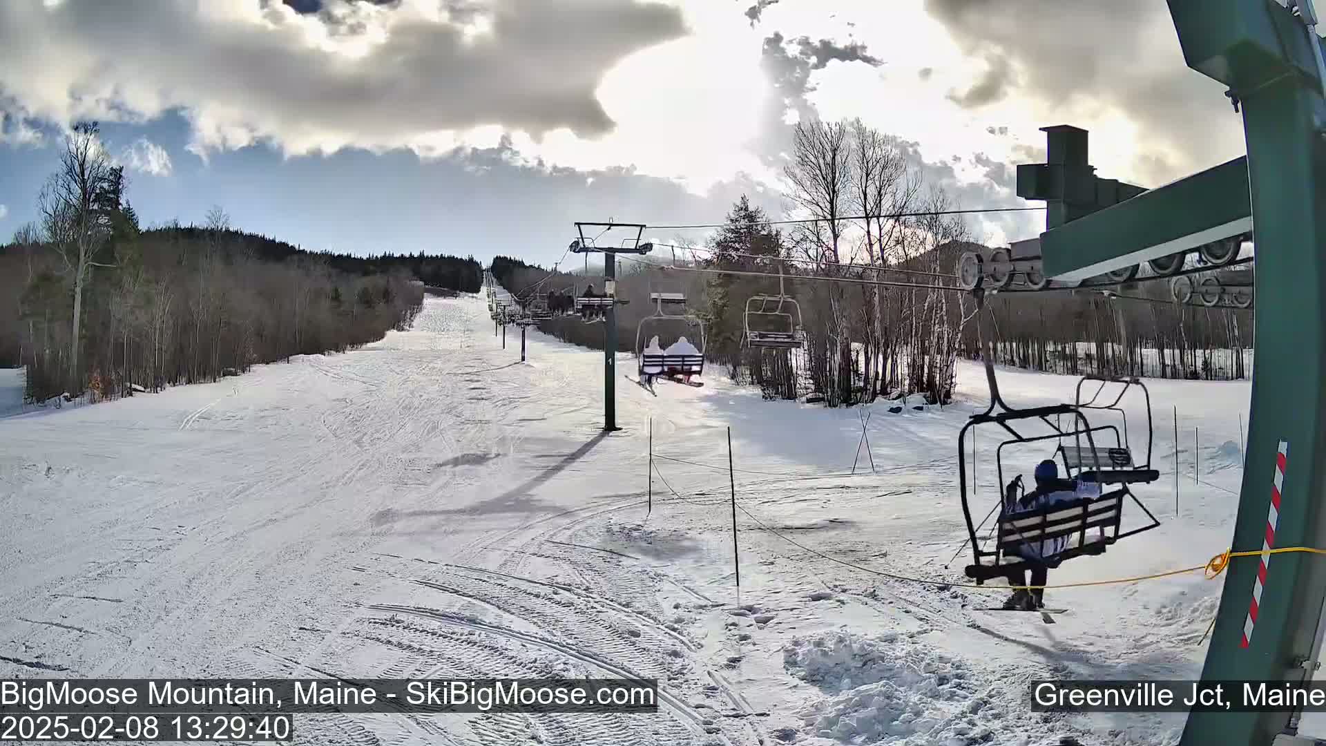 A snowy ski slope with a chairlift carrying skiers under a partly sunny sky.