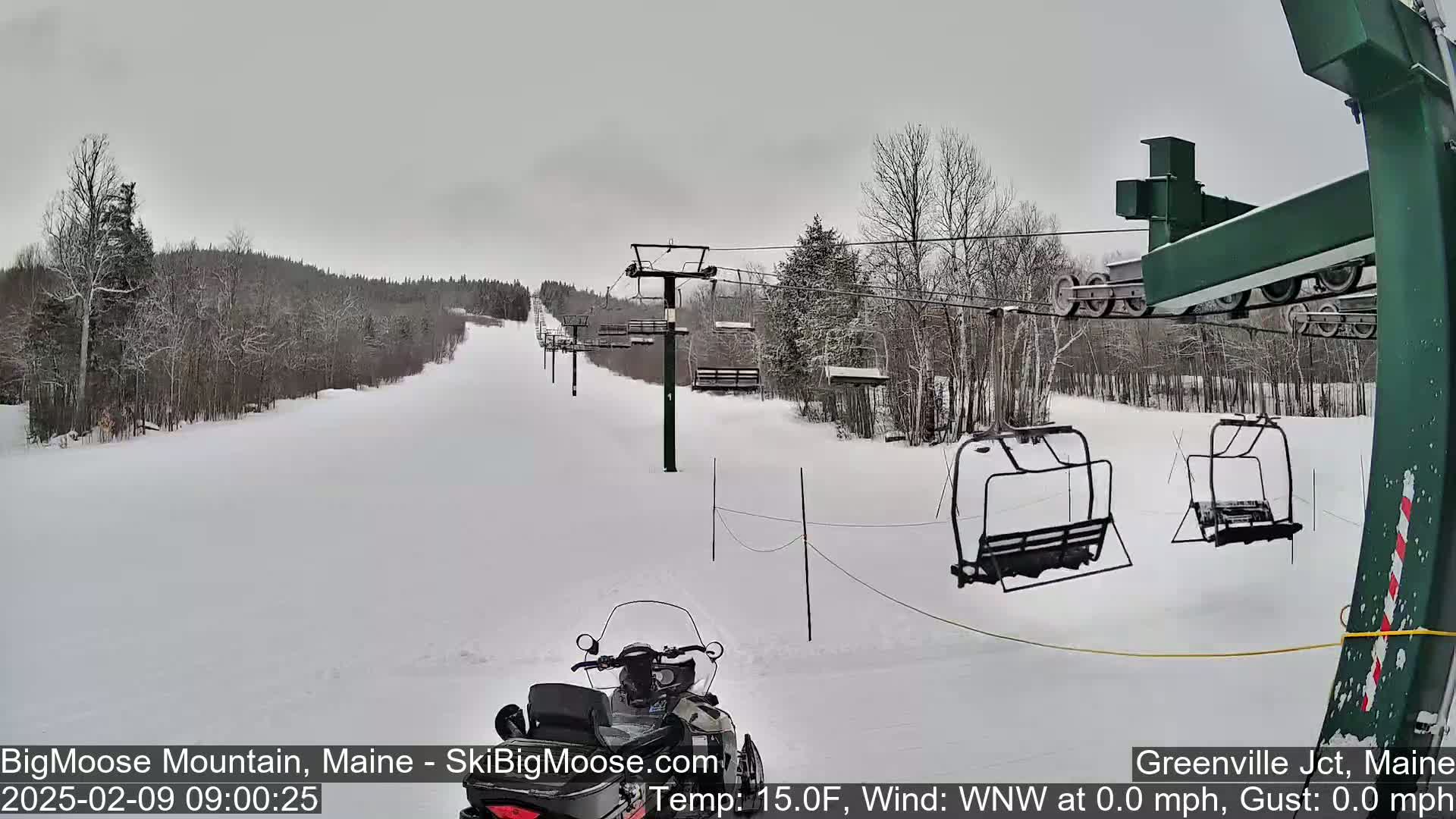 A snow-covered ski slope with a chairlift and a snowmobile, under an overcast sky.