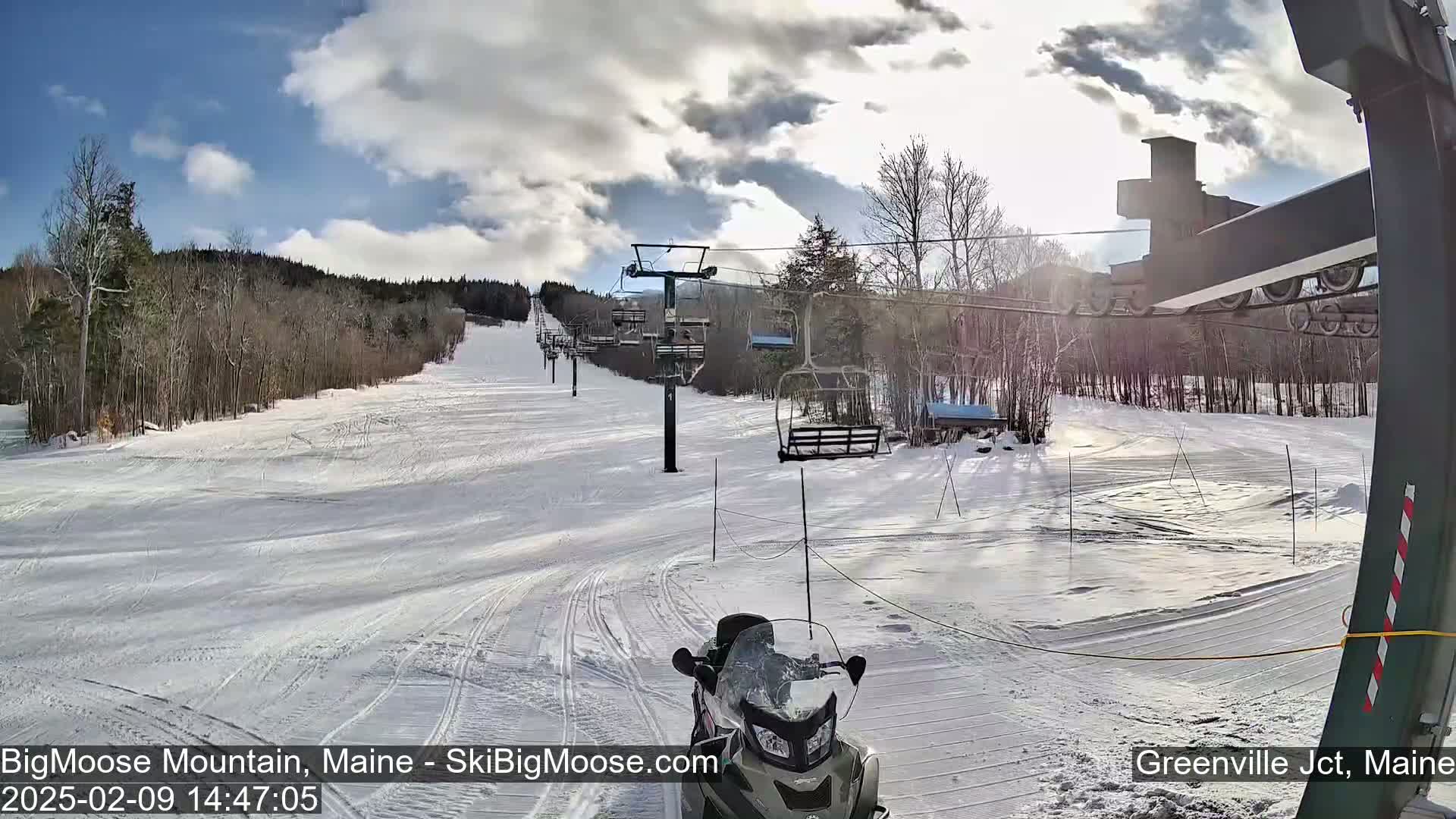 A snowmobile sits at the bottom of a snow-covered ski slope on a partly sunny day with some clouds.