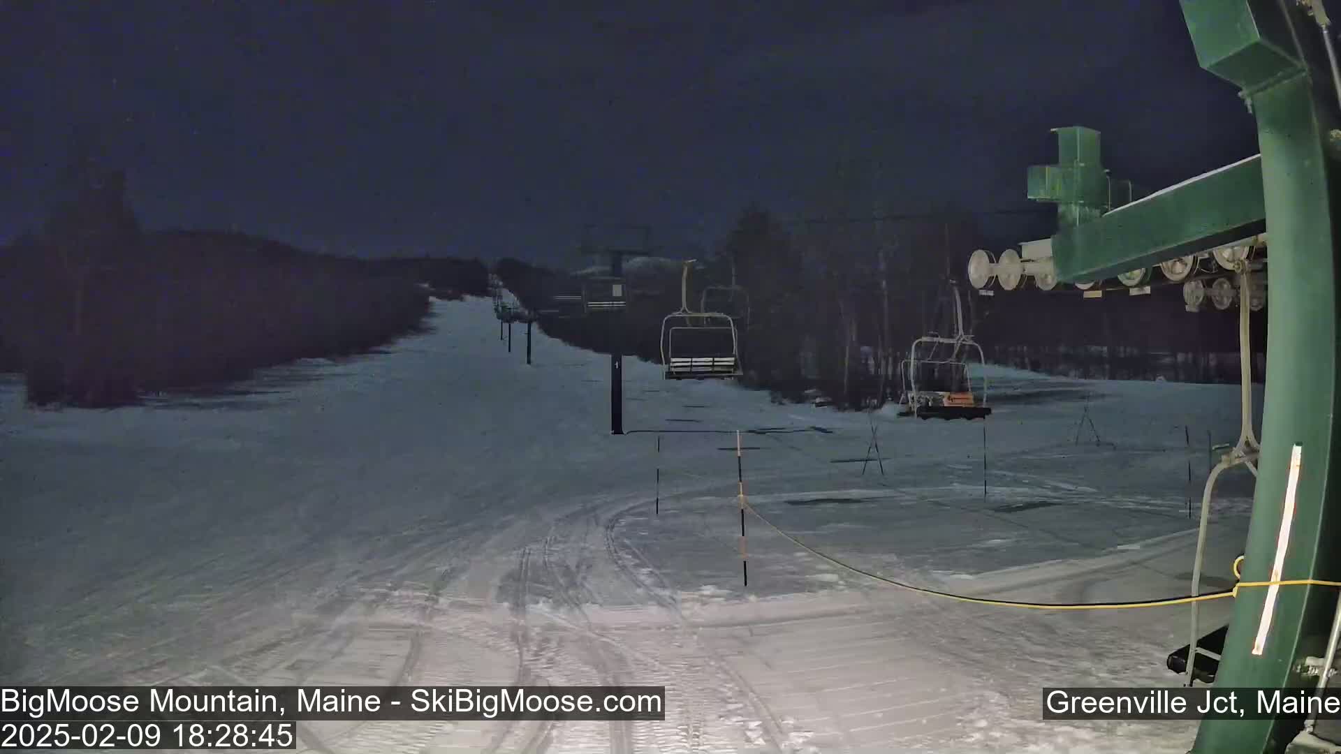 A snow-covered ski slope at night, illuminated by artificial lights, shows several empty chairlifts extending towards a dark treeline.
