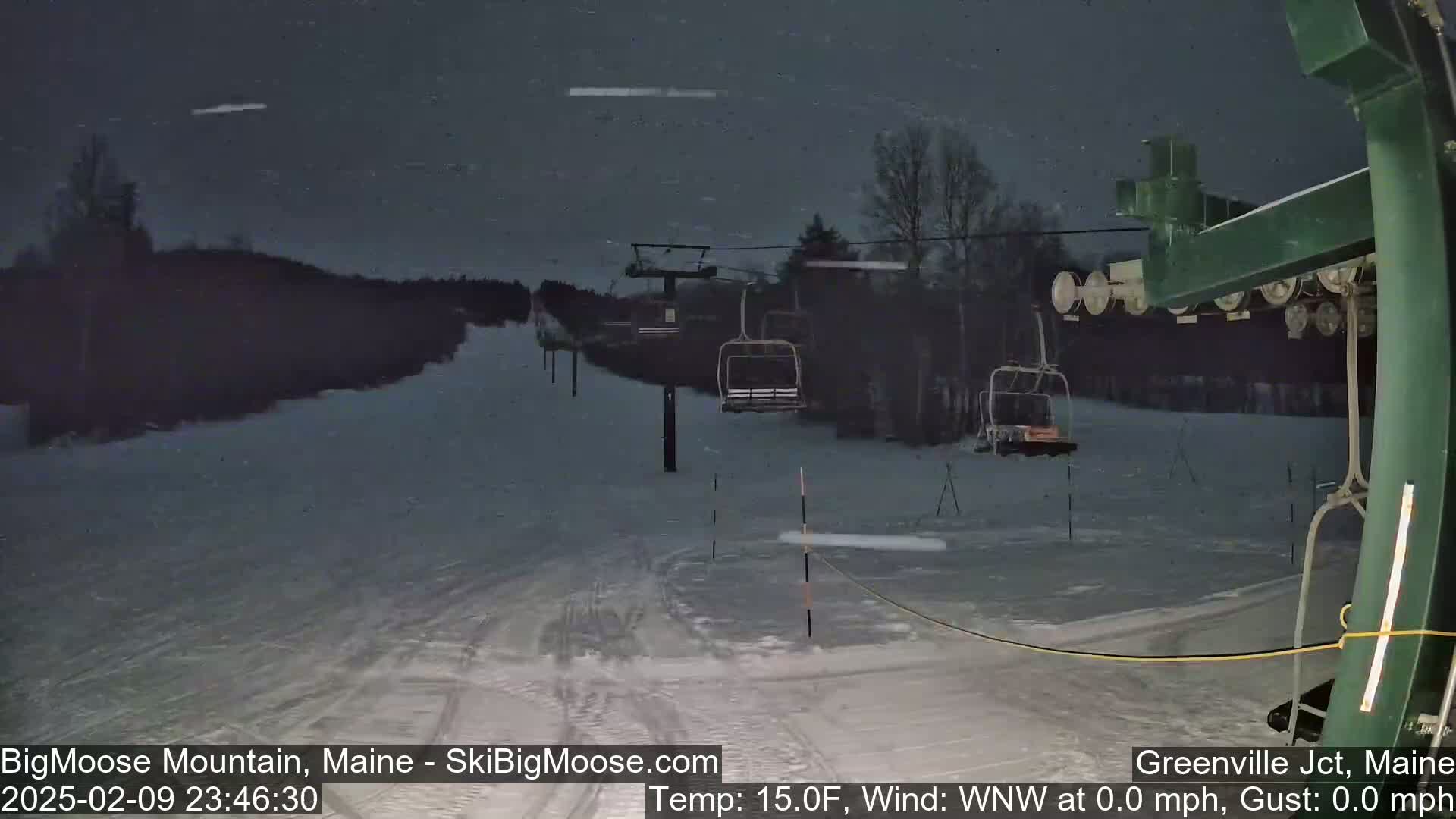 A nighttime view of a snow-covered ski slope with a chairlift and sparsely populated trees under a lightly snowing sky.