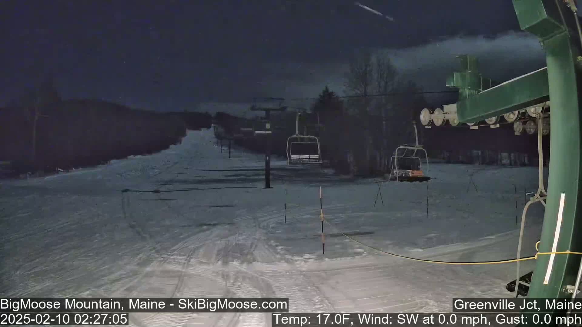 A nighttime view of a snow-covered ski slope with a chairlift and several empty chairs, under a partly cloudy sky.