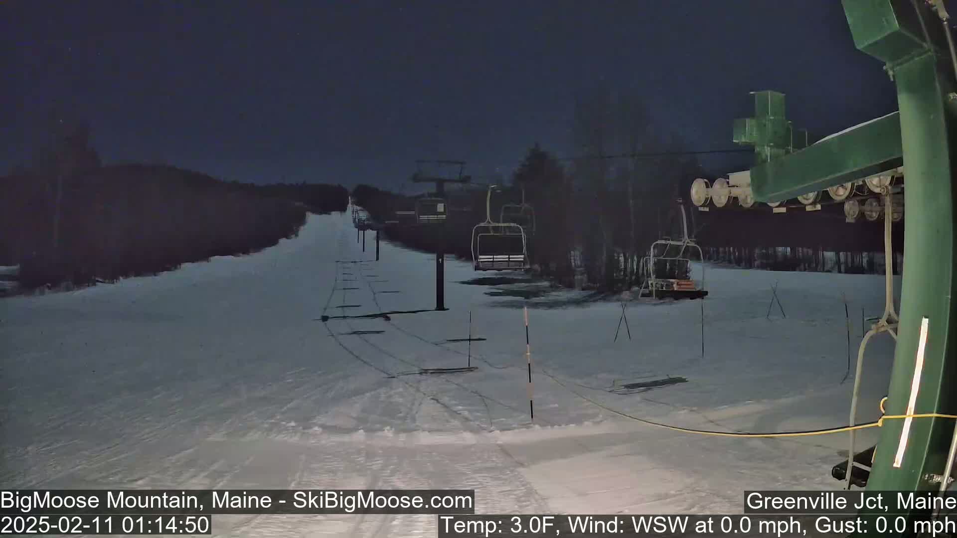 A snow-covered ski slope at night with a chairlift and several chairs visible under a dark sky.