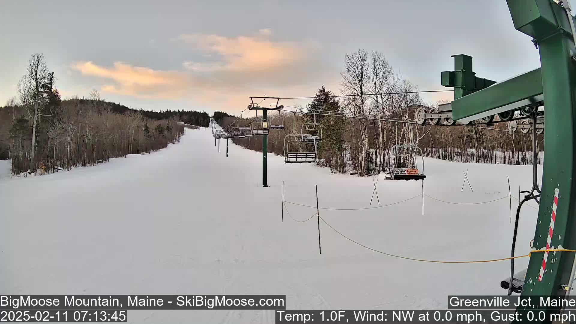 A snow-covered ski slope with a chairlift and bare trees under a partly cloudy sky.