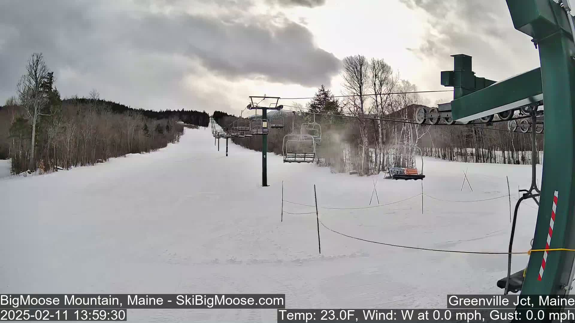 A snow-covered ski slope with a chairlift and sparsely wooded areas under an overcast sky.