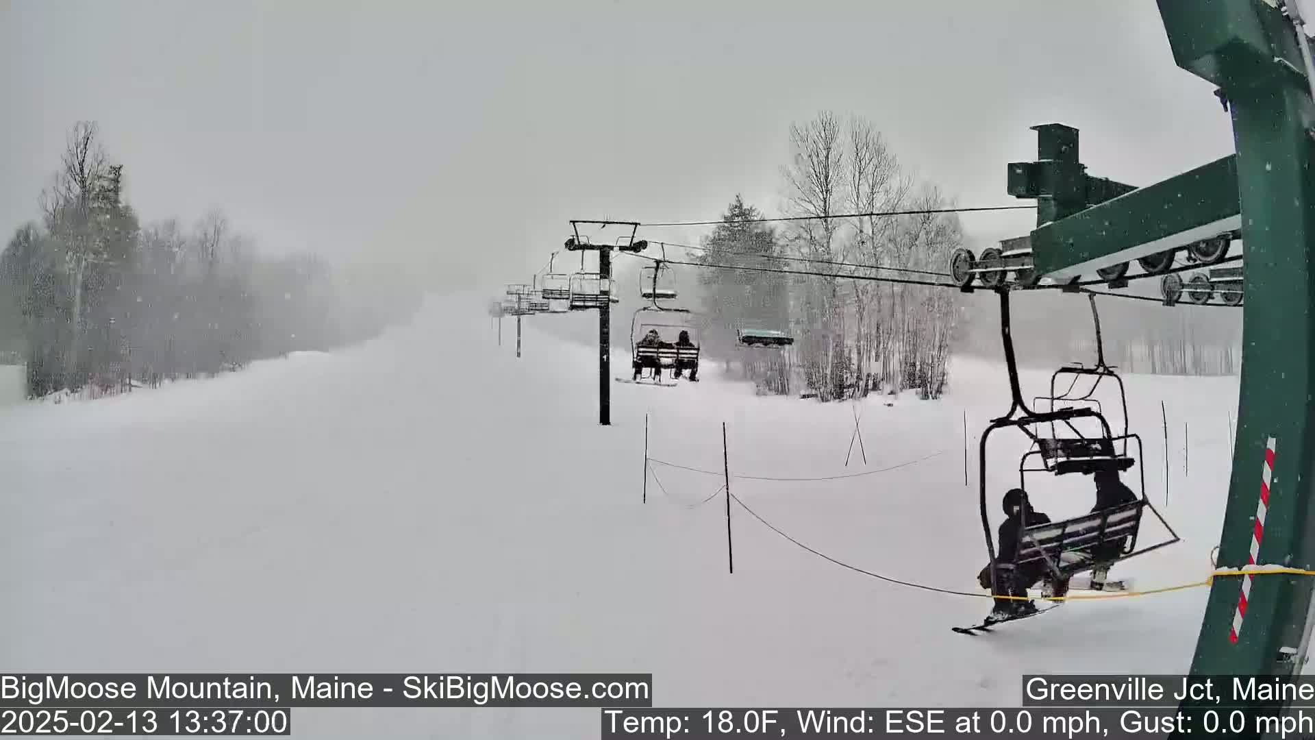 A snowy scene shows a ski lift carrying skiers up a snow-covered slope in heavy snowfall.